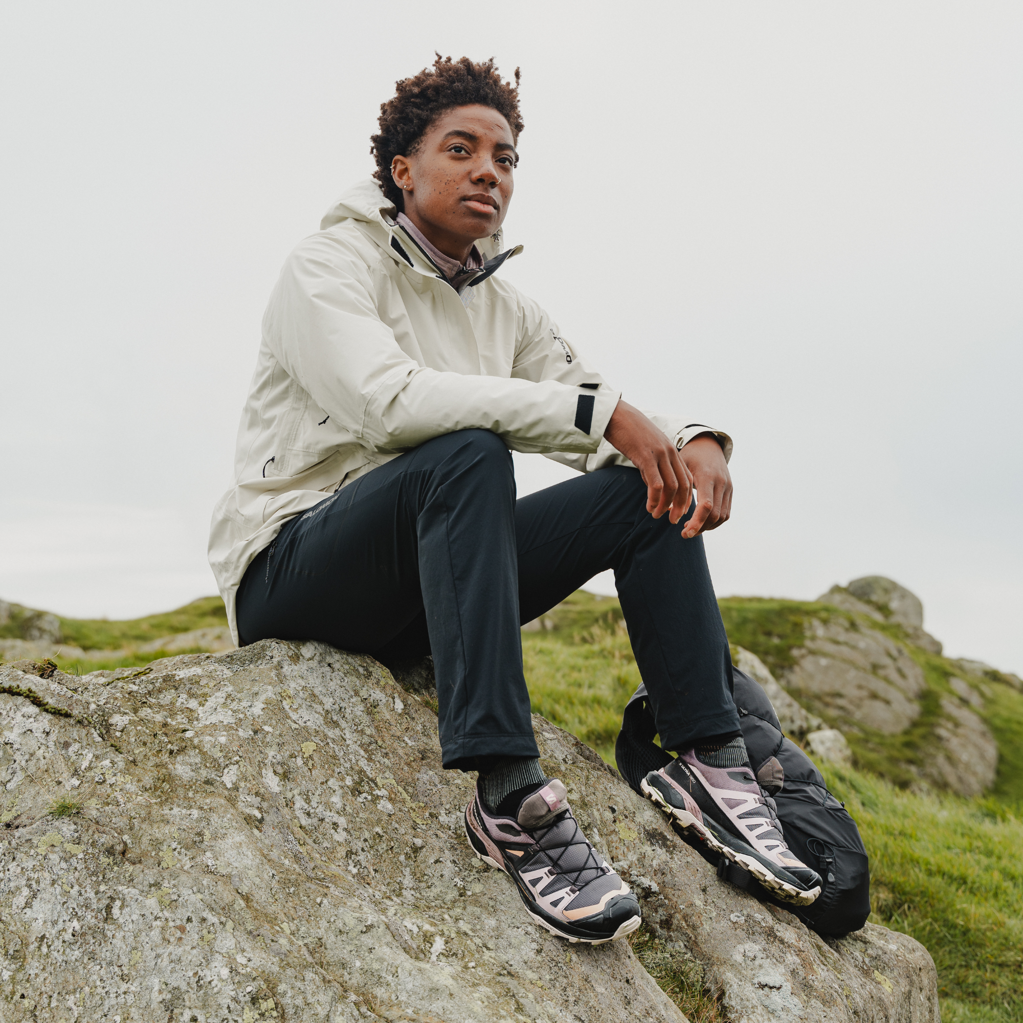 A woman sits on a large rock outdoors wearing a light beige hooded jacket, dark trousers, and Salomon Women's X Ultra 360 Gore-Tex Hiking Trail Shoes Plum Kitten Phantom Cork L47449400, with a black backpack beside her on a grassy hillside under an overcast sky.