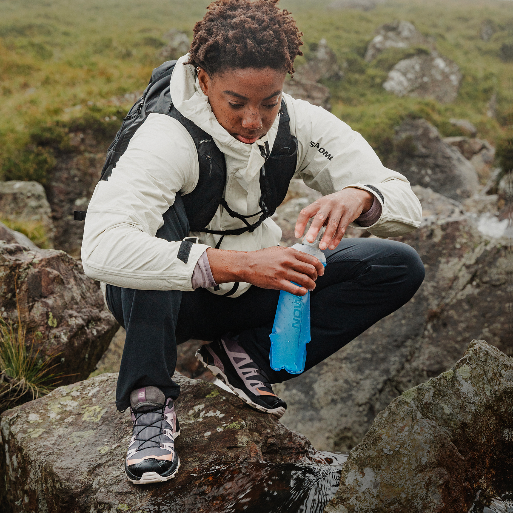 A person crouching outdoors on rocks, opening a blue water bottle while wearing a white Salomon jacket, black backpack, and dark trousers and Salomon Women's X Ultra 360 Gore-Tex Hiking Trail Shoes Plum Kitten Phantom Cork L47449400.