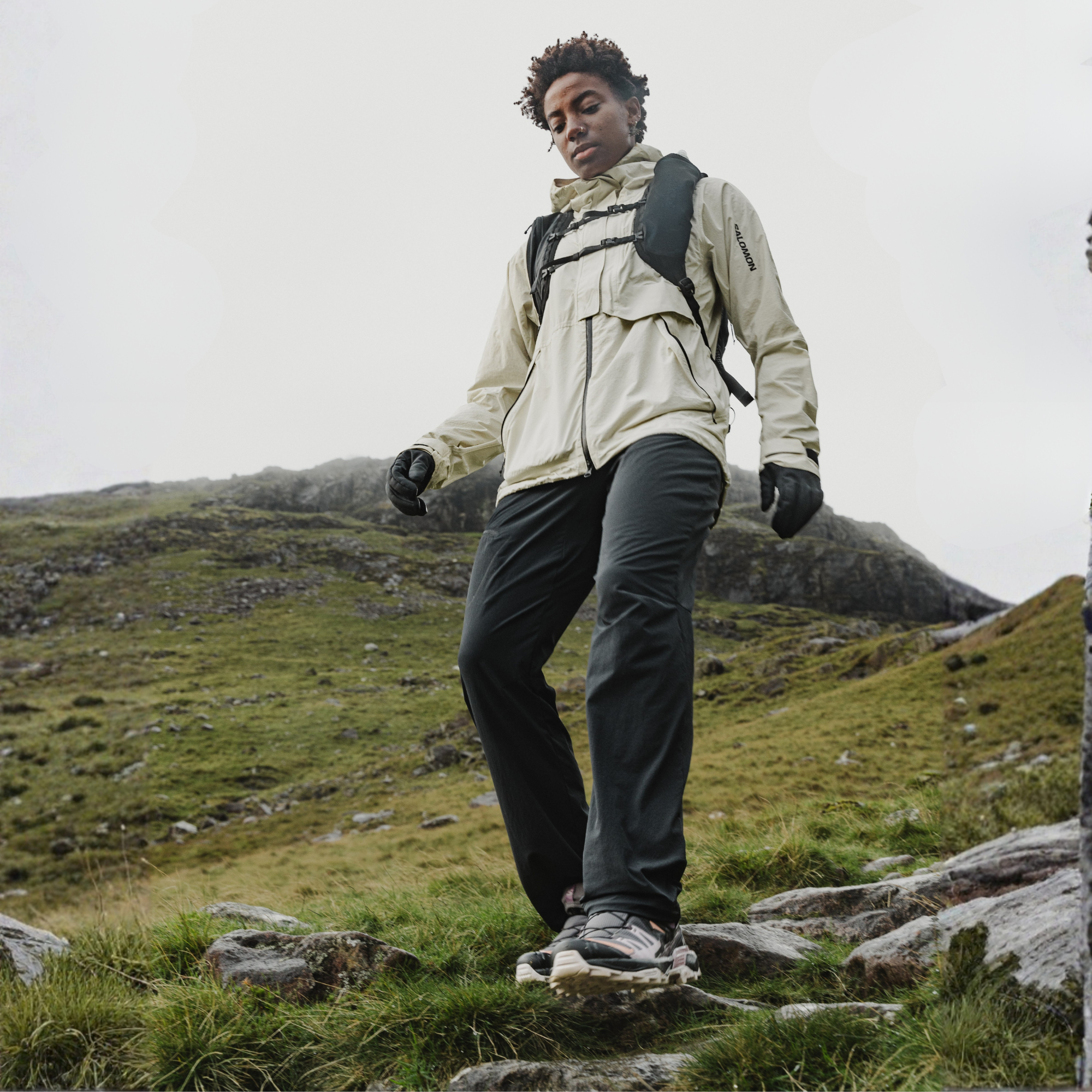 A woman hiking outdoors on a rocky grassy mountain slope, wearing a light beige technical jacket, dark grey hiking trousers, black gloves, Salomon Women's X Ultra 360 Gore-Tex Hiking Trail Shoes Plum Kitten Phantom Cork L47449400 and a black backpack.