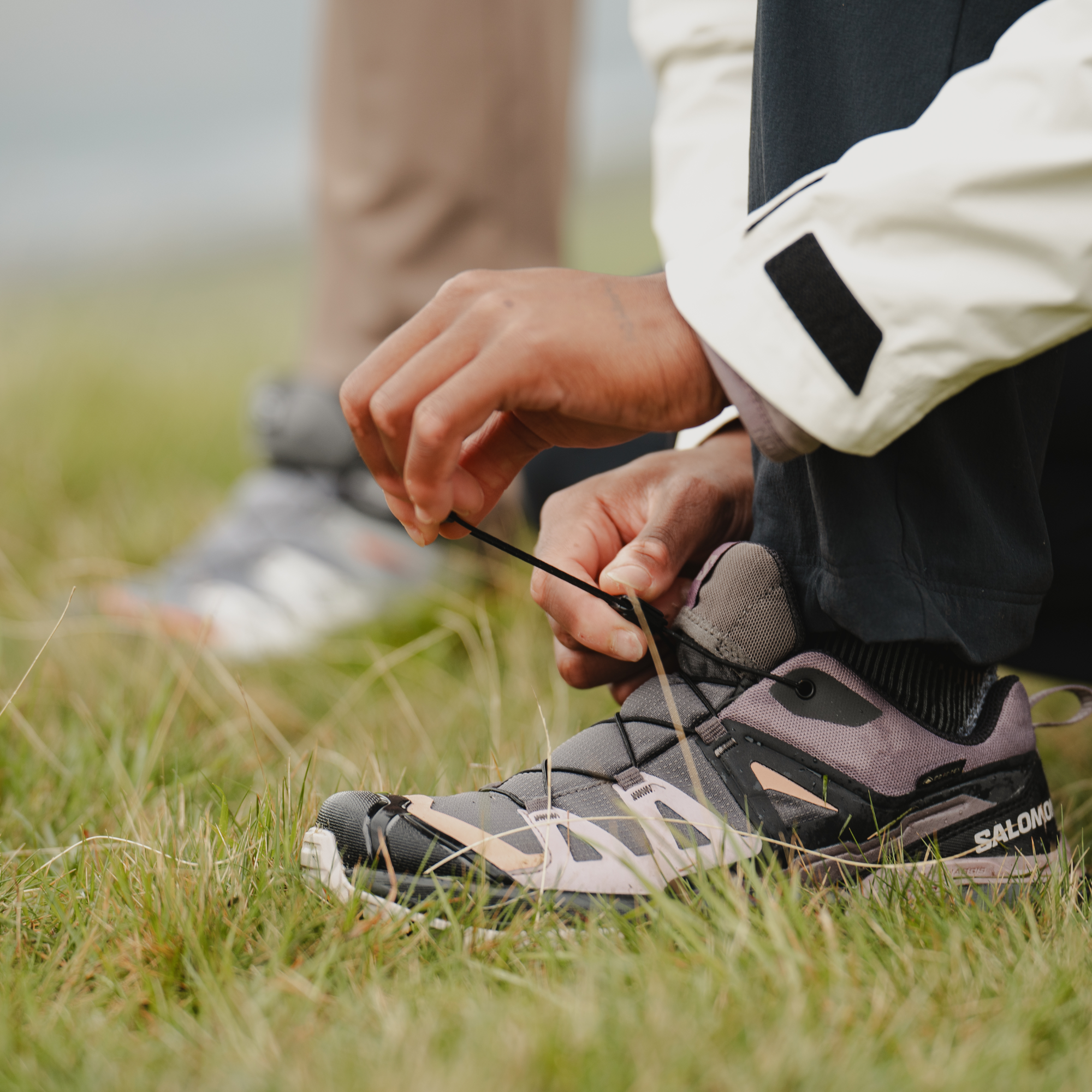 A close-up outdoor shot of a person tying the laces of Salomon Women's X Ultra 360 Gore-Tex Hiking Trail Shoes Plum Kitten Phantom Cork L47449400 while wearing dark trousers and a light-coloured jacket, with grass and another person's legs blurred in the background.