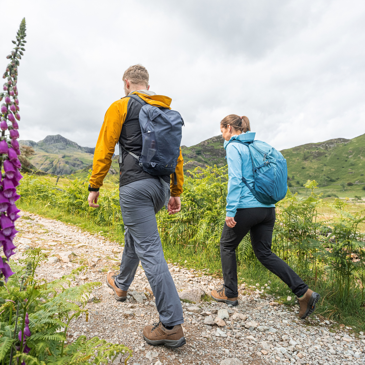 Two hikers walking on a gravel path in a mountainous outdoor setting, viewed from behind. The man wears a yellow and black jacket with a dark blue backpack and Scarpa Men's Cyrus 2 Gore-Tex Trail Shoes Brown Black 63326-200, and the woman wears a light blue jacket with a bright blue backpack.