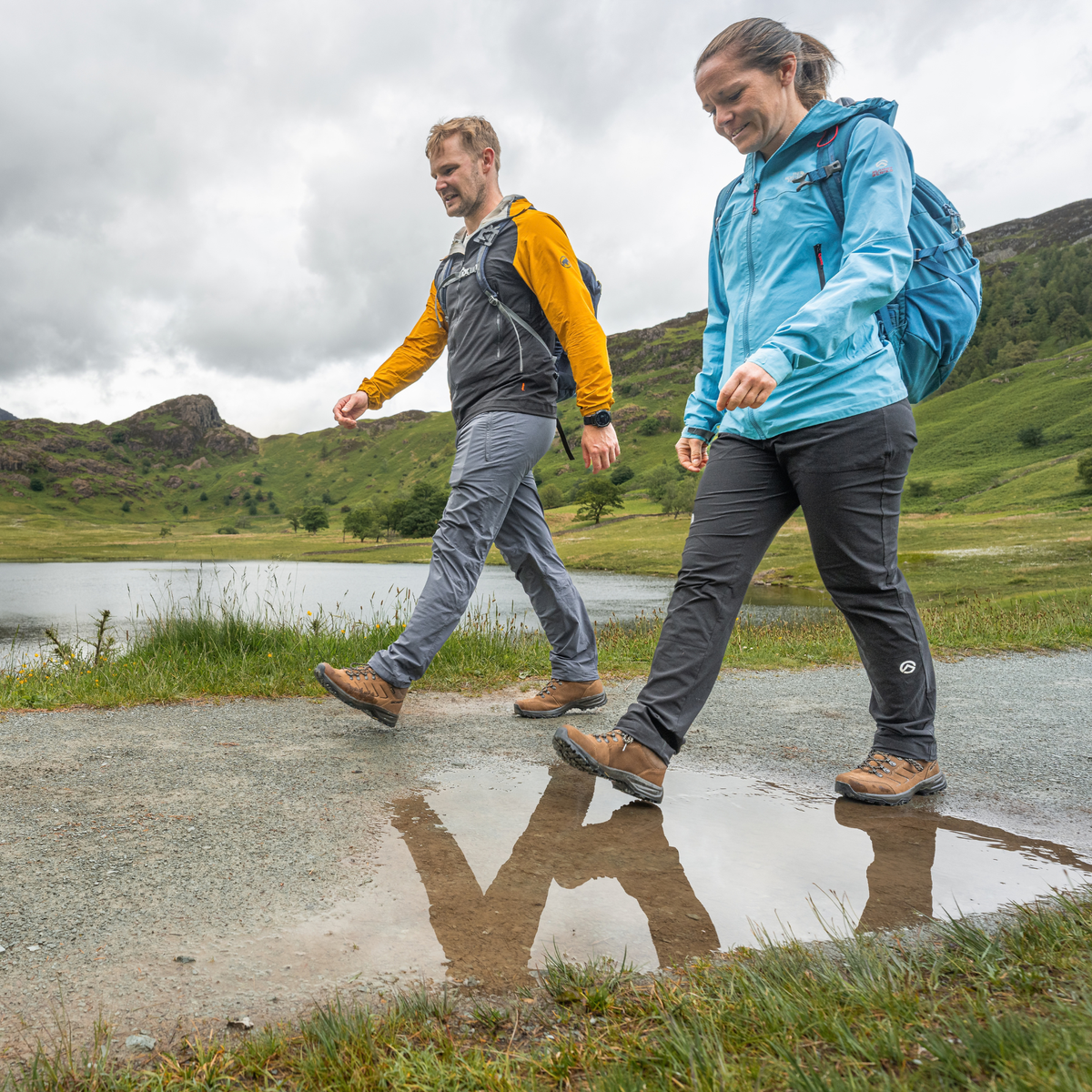 Two people walking outdoors on a gravel path near a lake, wearing waterproof jackets and trousers. The person in front wears a bright blue jacket and dark grey trousers, while the person behind wears a yellow-and-grey jacket, grey trousers and Scarpa Men's Cyrus 2 Gore-Tex Trail Shoes Brown Black 63326-200.