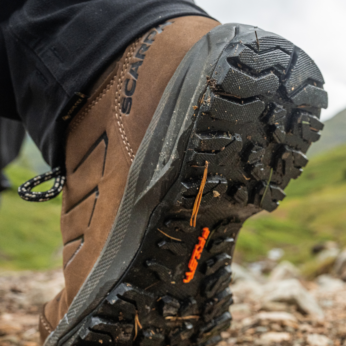 Close-up of the sole of a Scarpa Men's Cyrus 2 Gore-Tex Trail Shoes Brown Black 63326-200, worn with black trousers on a rocky outdoor trail.