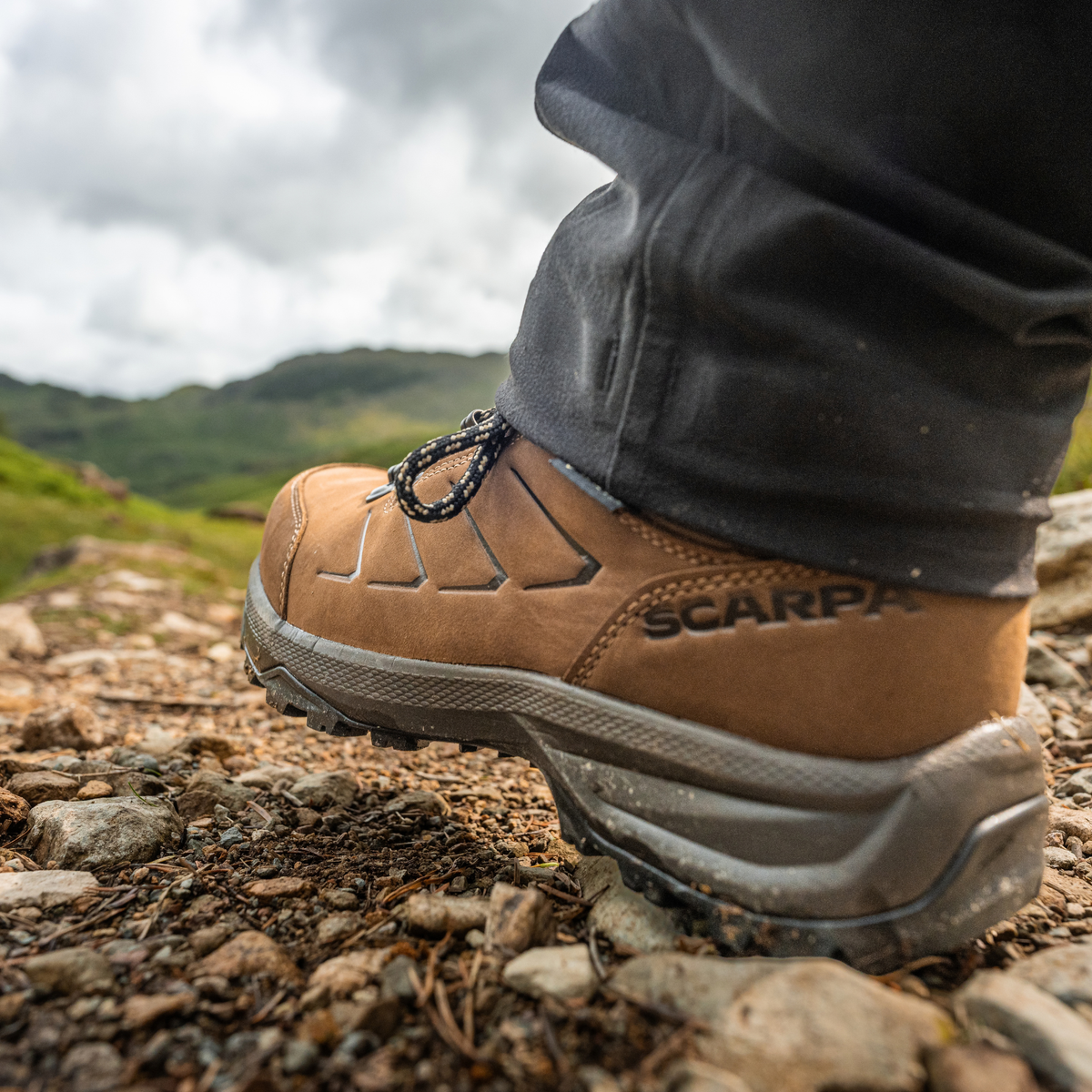A close-up shot of a person's foot wearing a Scarpa Men's Cyrus 2 Gore-Tex Trail Shoes Brown Black 63326-200 with dark grey trousers, standing on a rocky dirt path with a blurred mountain landscape in the background.