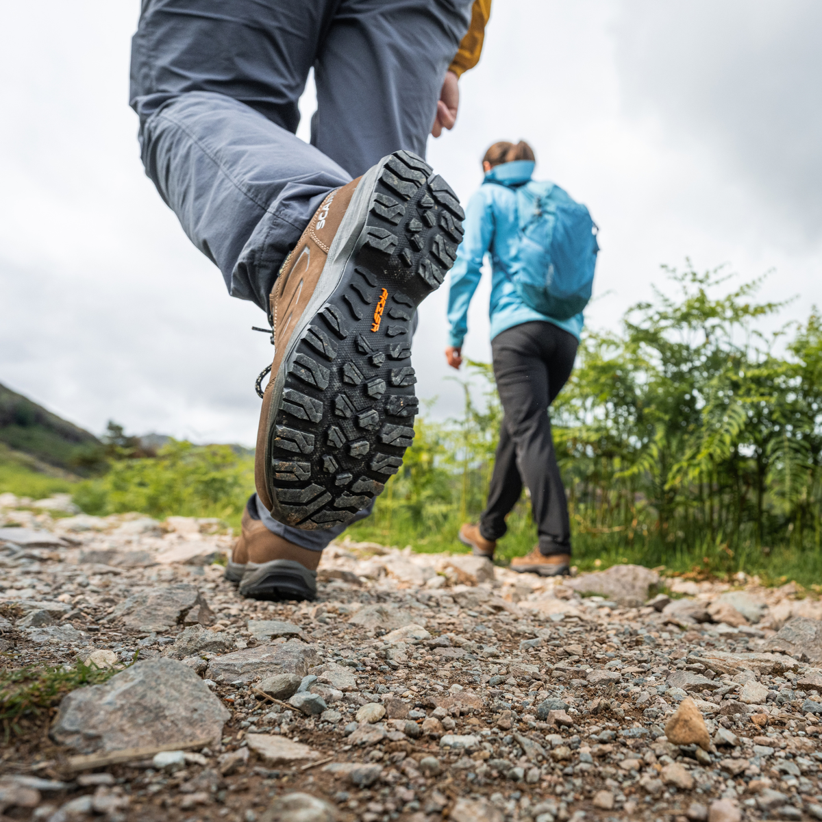 A close-up of a person hiking on a rocky trail, showing the treaded sole of a Scarpa Men's Cyrus 2 Gore-Tex Trail Shoes Brown Black 63326-200. Another person in a blue jacket walks in the background under an overcast sky.