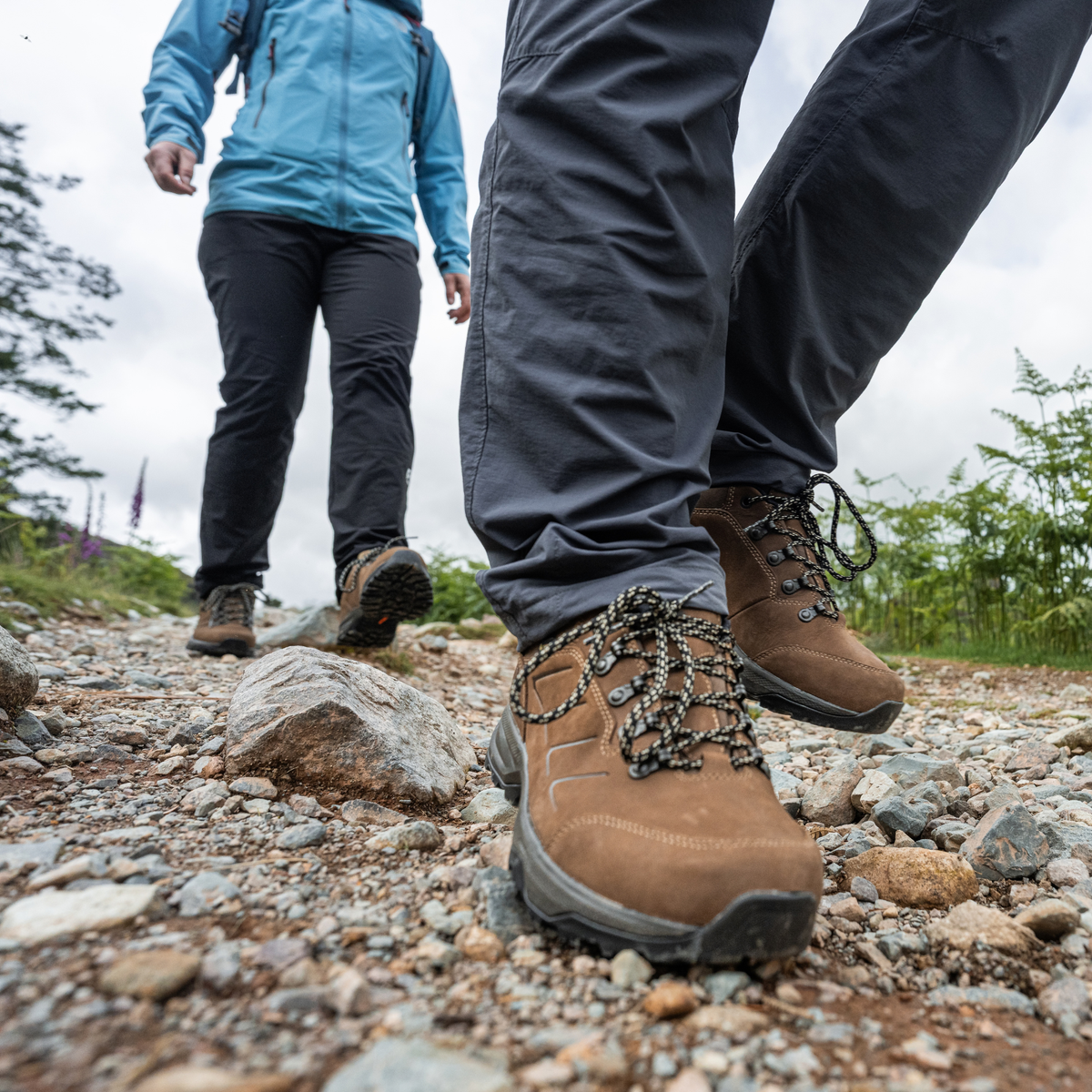 Close-up of a person hiking on a rocky trail wearing Scarpa Men's Cyrus 2 Gore-Tex Trail Shoes Brown Black 63326-200, dark grey trousers with another hiker visible in the background.