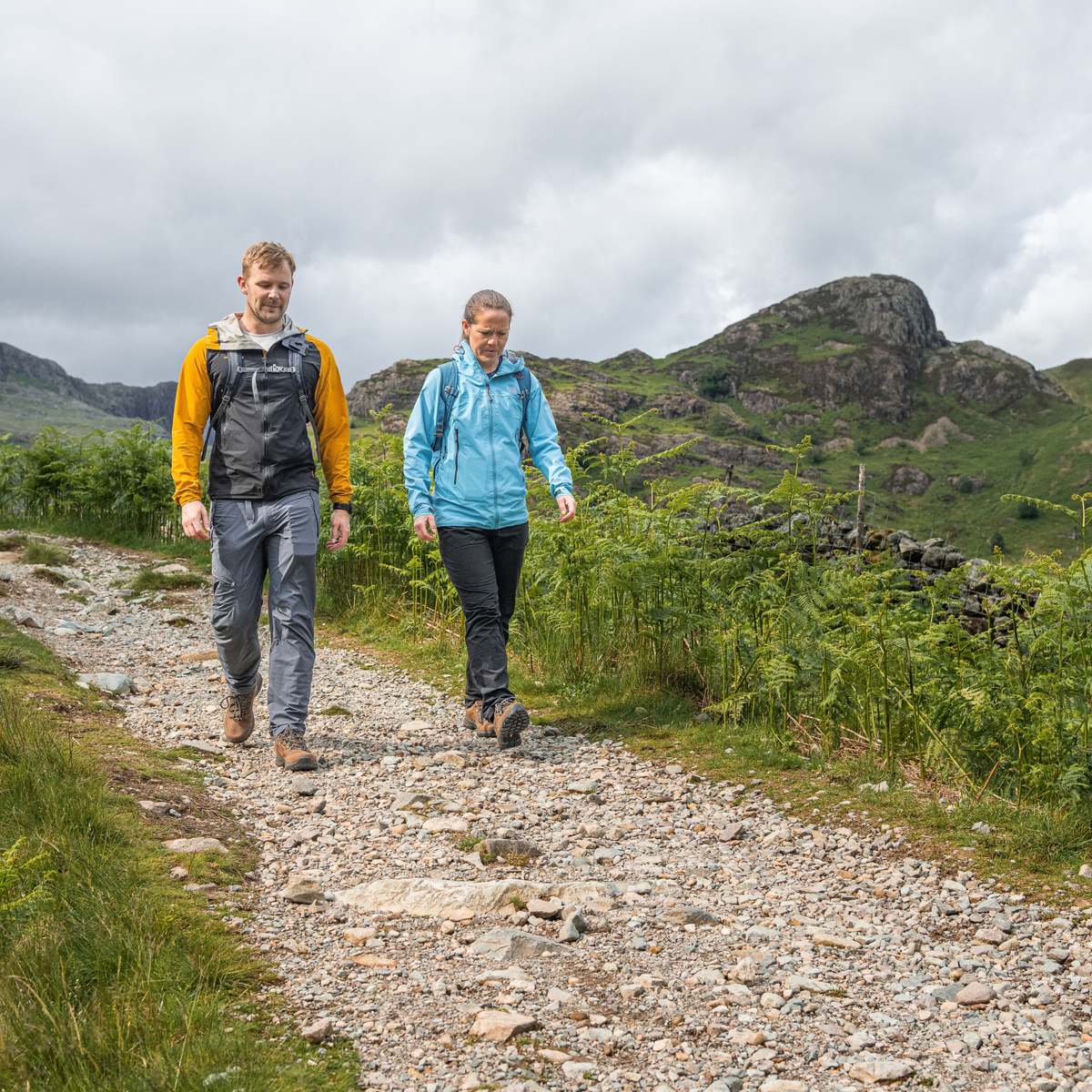 Two people hiking on a rocky path in a grassy mountain landscape under a cloudy sky. Man wearing Scarpa Men's Cyrus 2 Gore-Tex Trail Shoes Brown Black 63326-200.