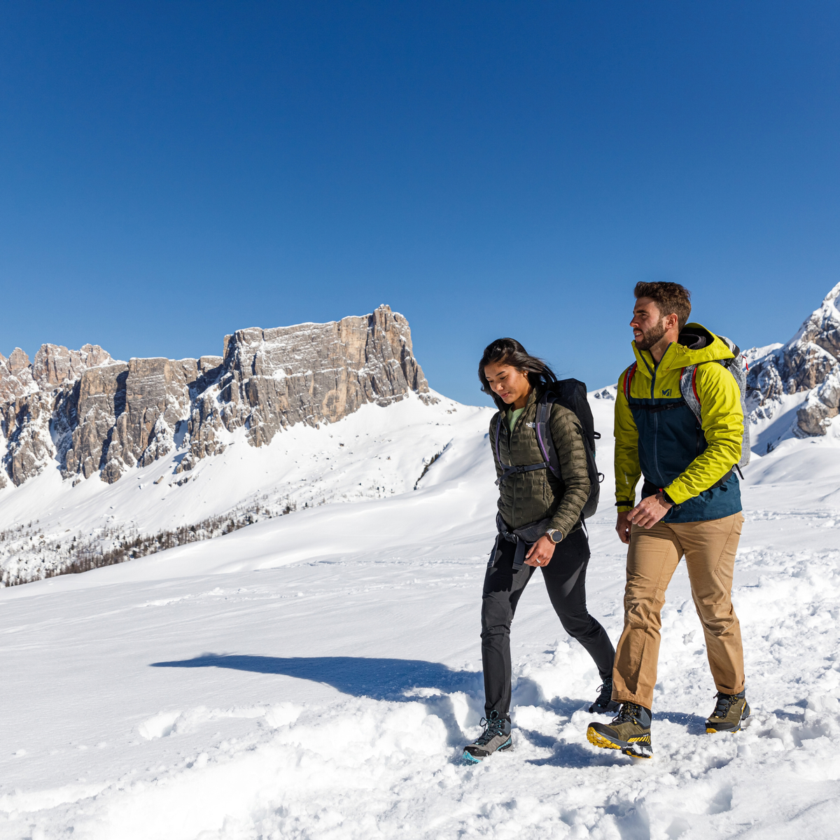 Two hikers walking in a snowy mountain landscape under a clear blue sky, with rocky peaks in the background. One wears a dark olive puffer jacket and black trousers and Scarpa Women's Rush TRK Gore-Tex Hillwalking Boots Mid Grey Aqua 63143-202, and the other wears a bright yellow and navy jacket with tan trousers.