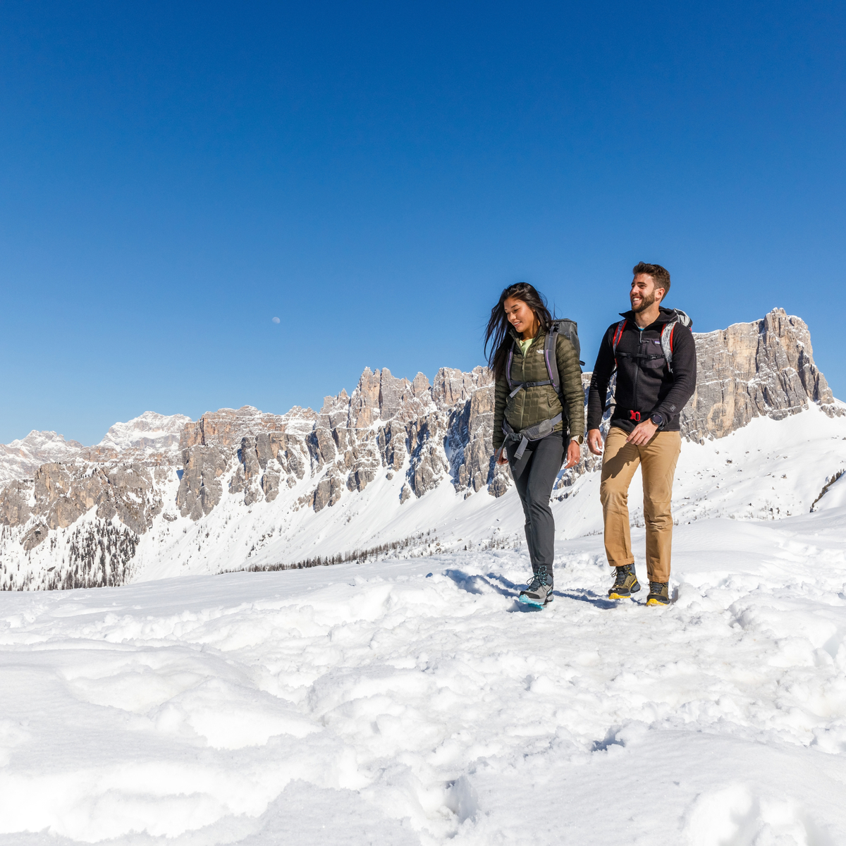 A man and a woman hiking together on a snowy mountain ridge with dramatic rocky cliffs and a clear blue sky in the background. Woman wearing Scarpa Women's Rush TRK Gore-Tex Hillwalking Boots Mid Grey Aqua 63143-202.