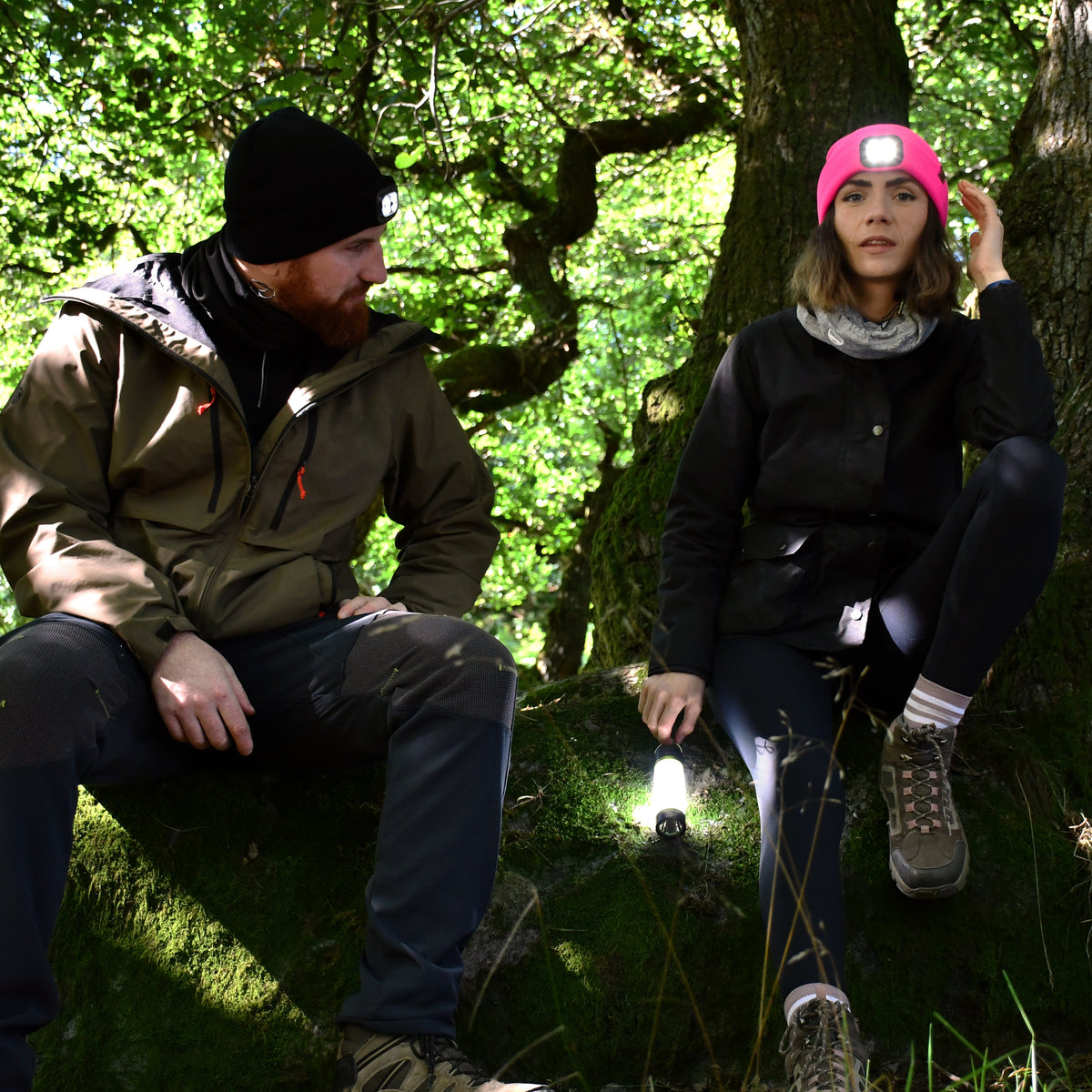 Two people sitting in a forested area with one wearing a Six Peaks Unisex LED Lighted Beanie Hat Pink SIH0004.