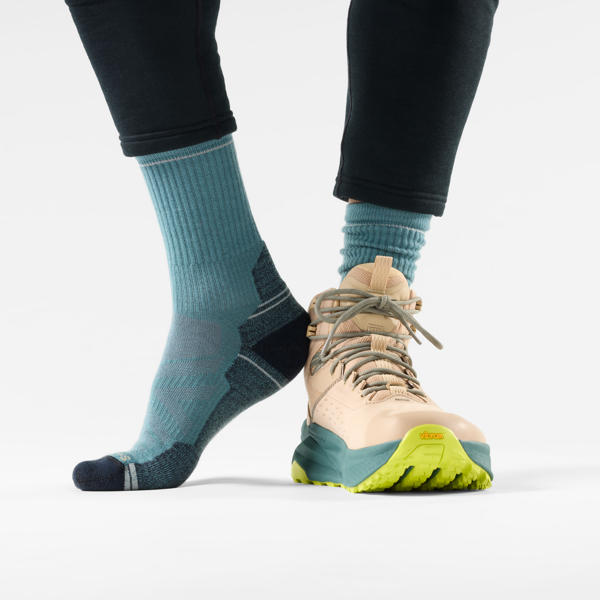 A close-up of a person's lower legs wearing Smartwool Men's Performance Hike Light Cushion Merino Blend Crew Socks Dusty Teal Q65 SW001614 with a salomon pink hiking boot featuring a bright yellow-green sole, set against a plain light grey background.