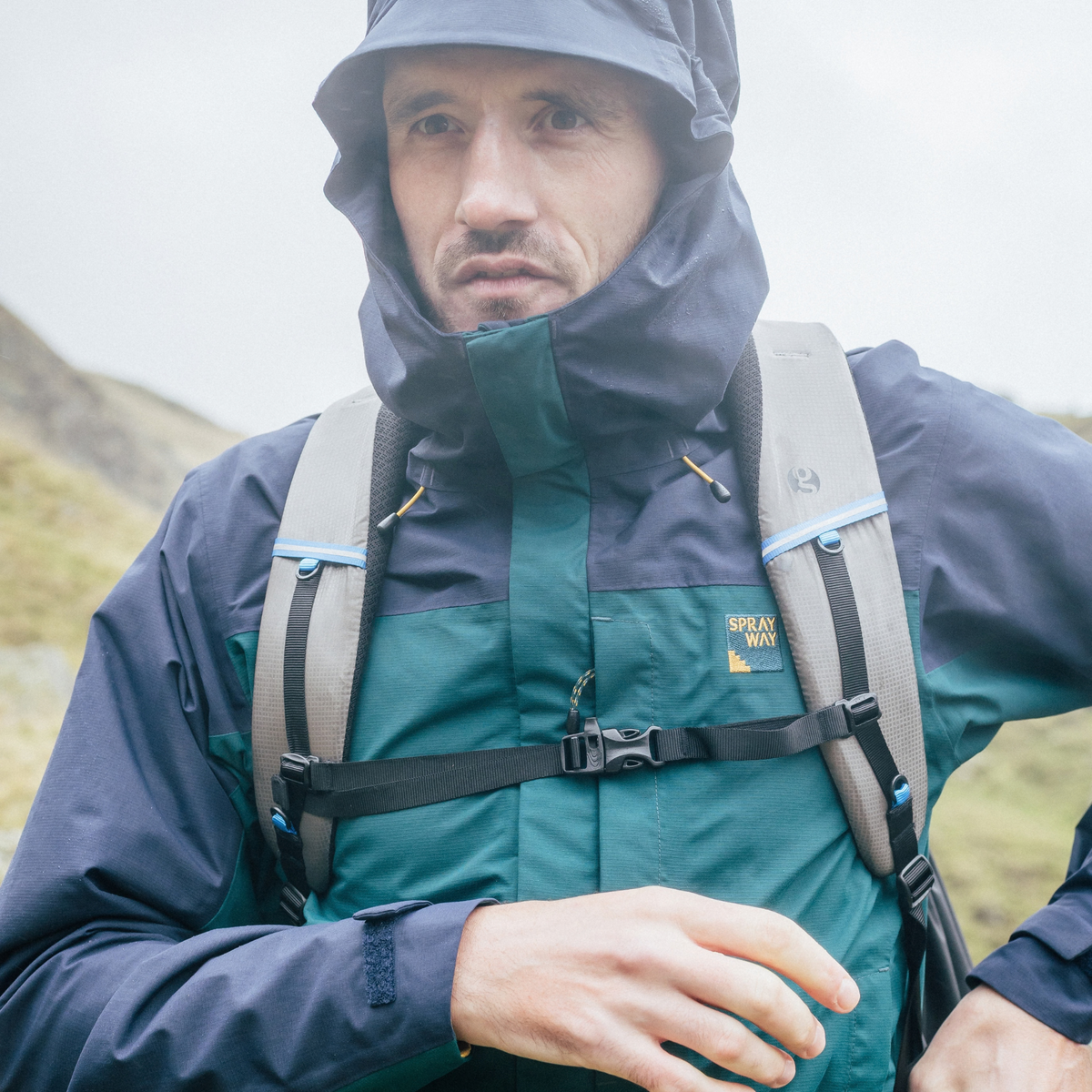 A man wearing a Sprayway Men's Maxen 2L Gore-Tex Waterproof Jacket SP-01754 Dark Sea Blazer SP-008873, a grey backpack, and a chest strap in an outdoor mountain setting.