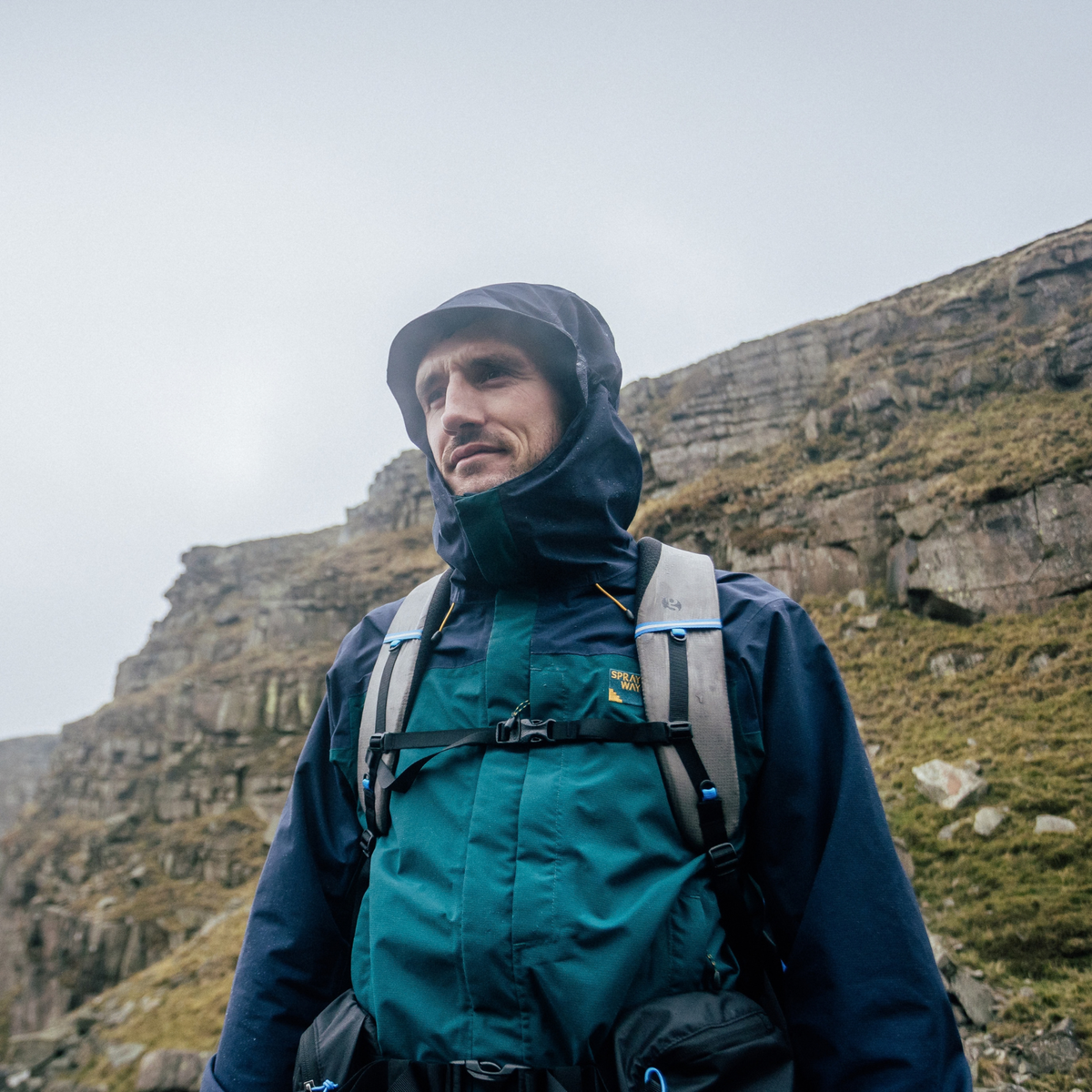 A man wearing aSprayway Men's Maxen 2L Gore-Tex Waterproof Jacket SP-01754 Dark Sea Blazer SP-008873 with a backpack stands outdoors against a rocky cliffside background under an overcast sky.