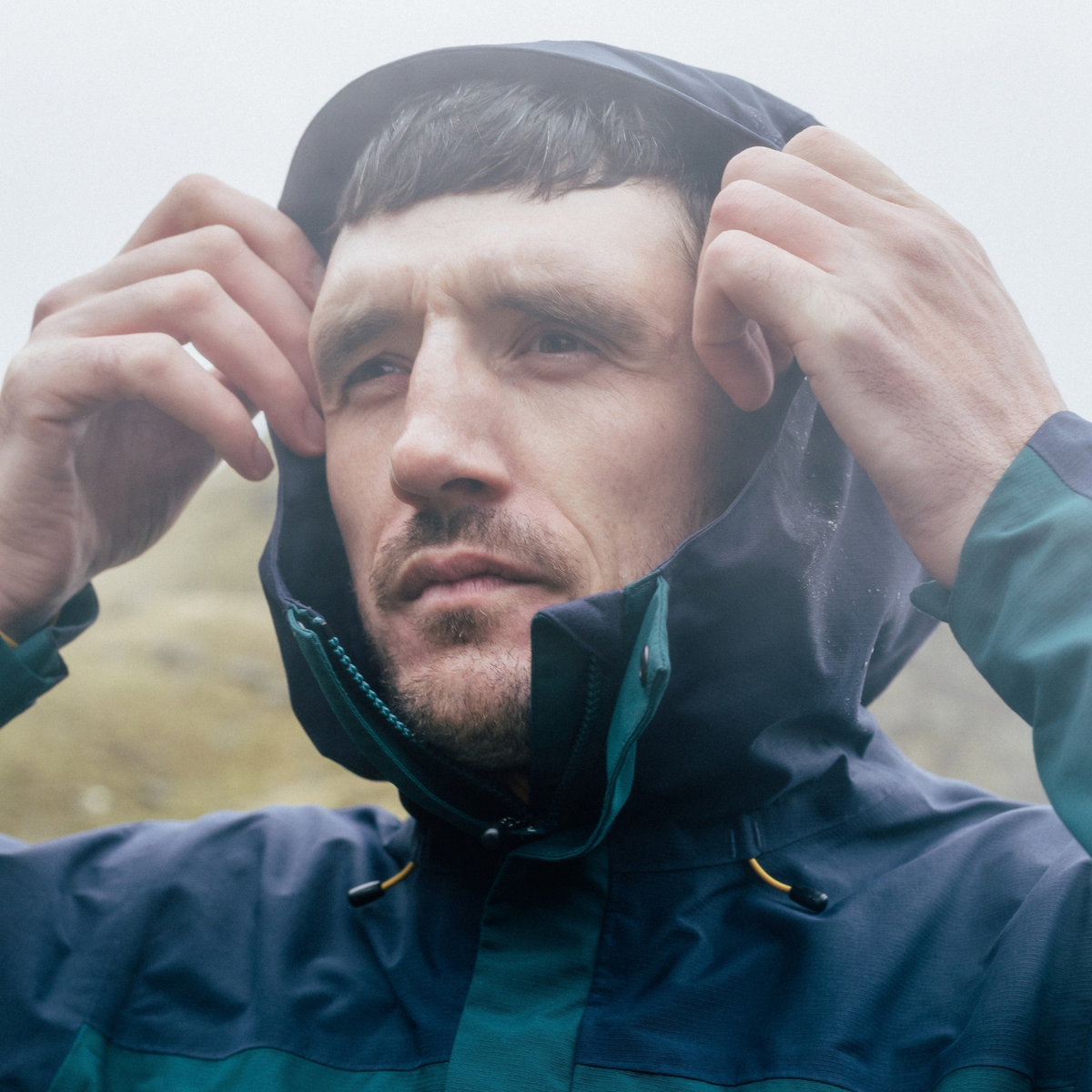 A man wearing a Sprayway Men's Maxen 2L Gore-Tex Waterproof Jacket SP-01754 Dark Sea Blazer SP-008873, adjusting the hood over his head in a misty outdoor setting.