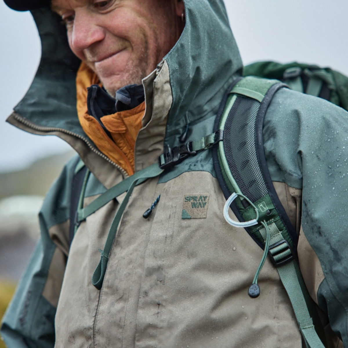 A close-up of a man wearing a Sprayway Men's Reaction 2L Gore-Tex Jacket SP-01723 Brownstone Bonsai SP-008036, and a hiking backpack strap over his shoulder against an overcast outdoor background.
