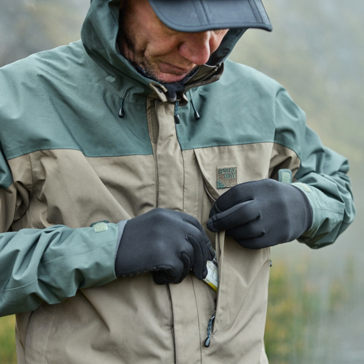 A man wearing a Sprayway Men's Reaction 2L Gore-Tex Jacket SP-01723 Brownstone Bonsai SP-008036, black gloves, and a dark cap, looking down while opening a pocket on his jacket. The background is a soft, blurred outdoor landscape.