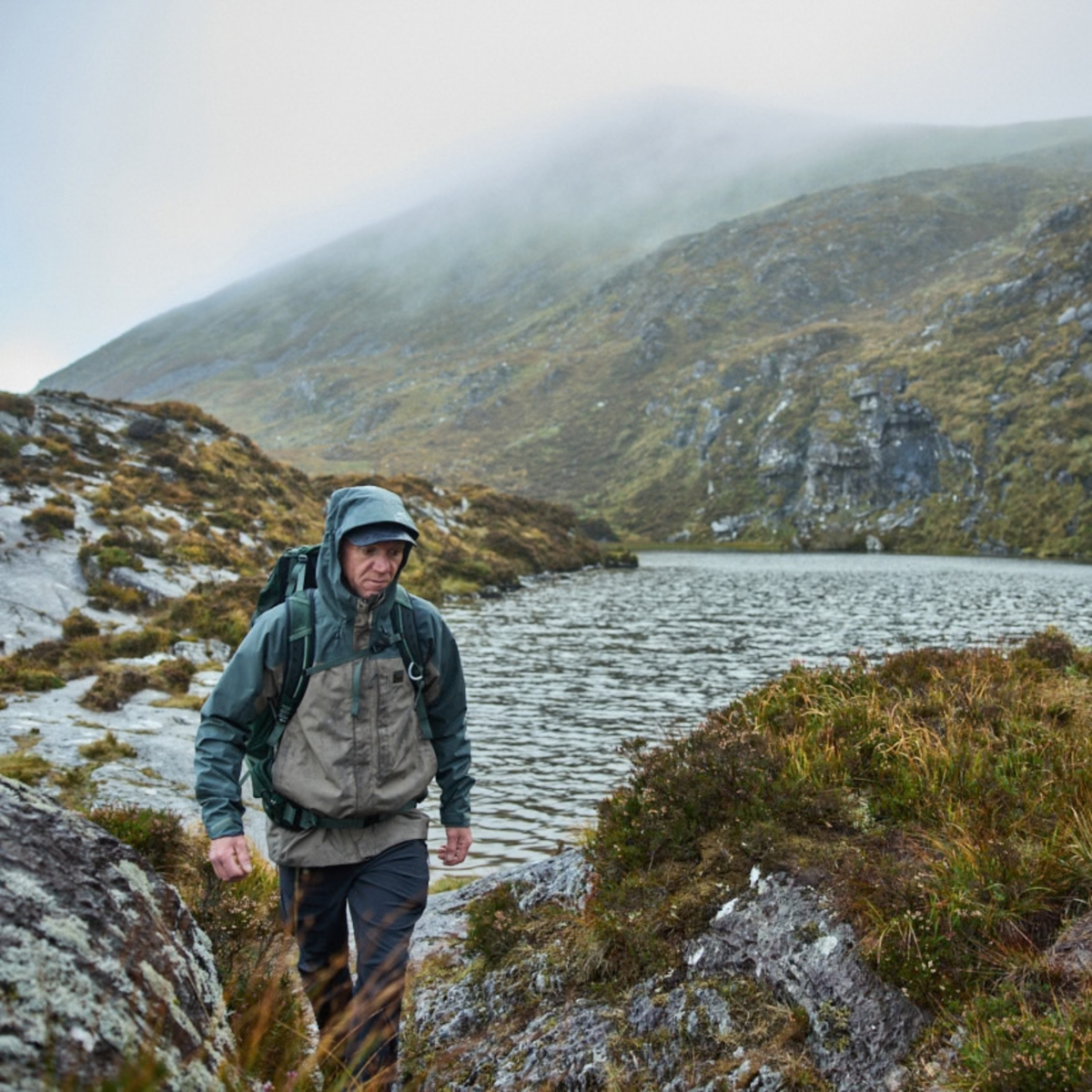 A man hiking in a rugged mountain landscape with a lake and misty hills in the background, wearing a Sprayway Men's Reaction 2L Gore-Tex Jacket SP-01723 Brownstone Bonsai SP-008036 and a backpack.