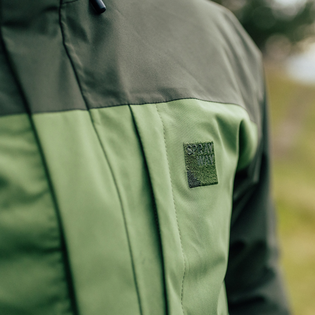 Close up of man wearing Sprayway Men's Santiago InterActive Waterproof Jacket SP-01753 Cypress Forest SP-008867 showing the Sprayway logo on chest with a blurry outdoor background
