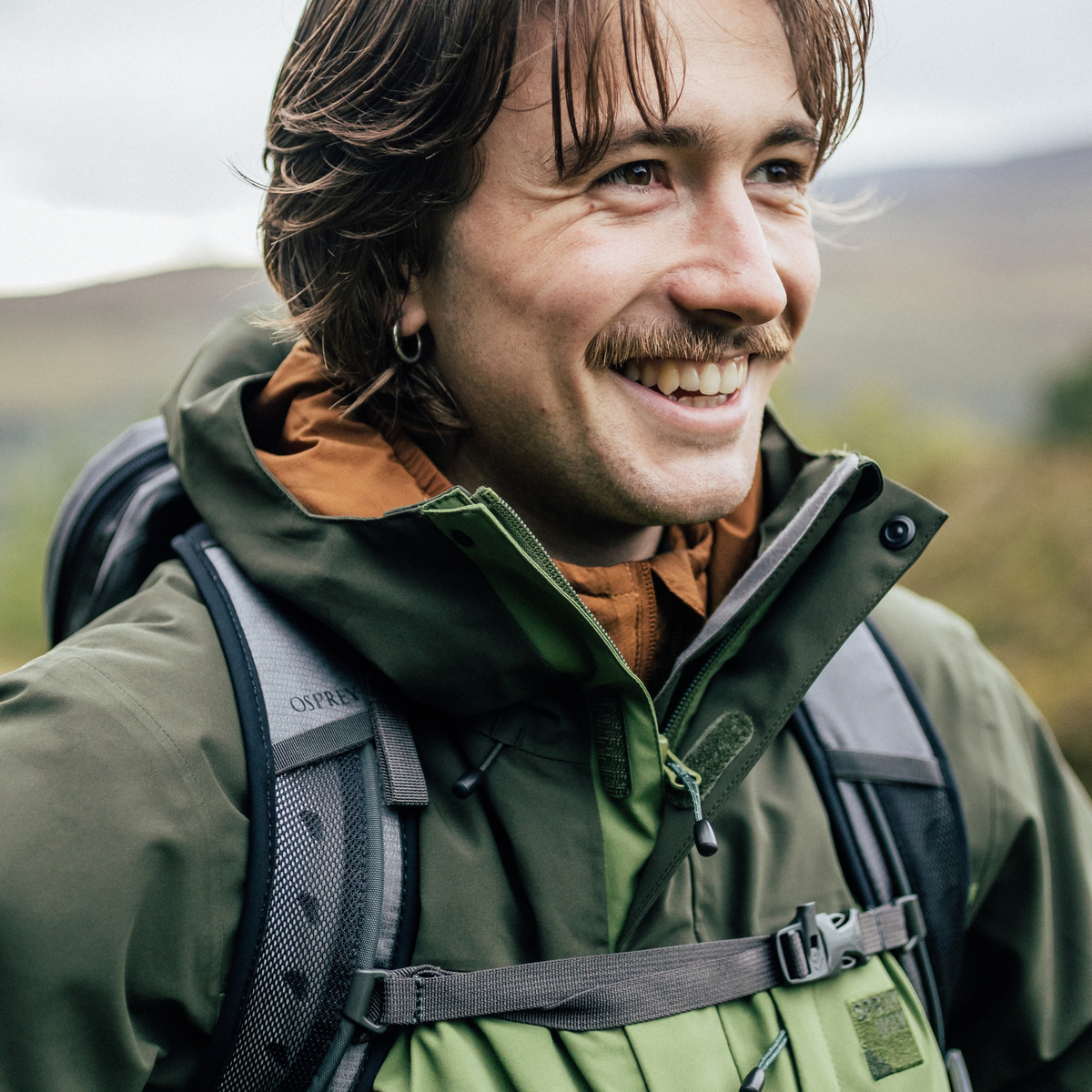 Close up of a man wearing Sprayway Men's Santiago InterActive Waterproof Jacket SP-01753 Cypress Forest SP-008867 smiling with a backpack on and a blurred outdoor mountain background