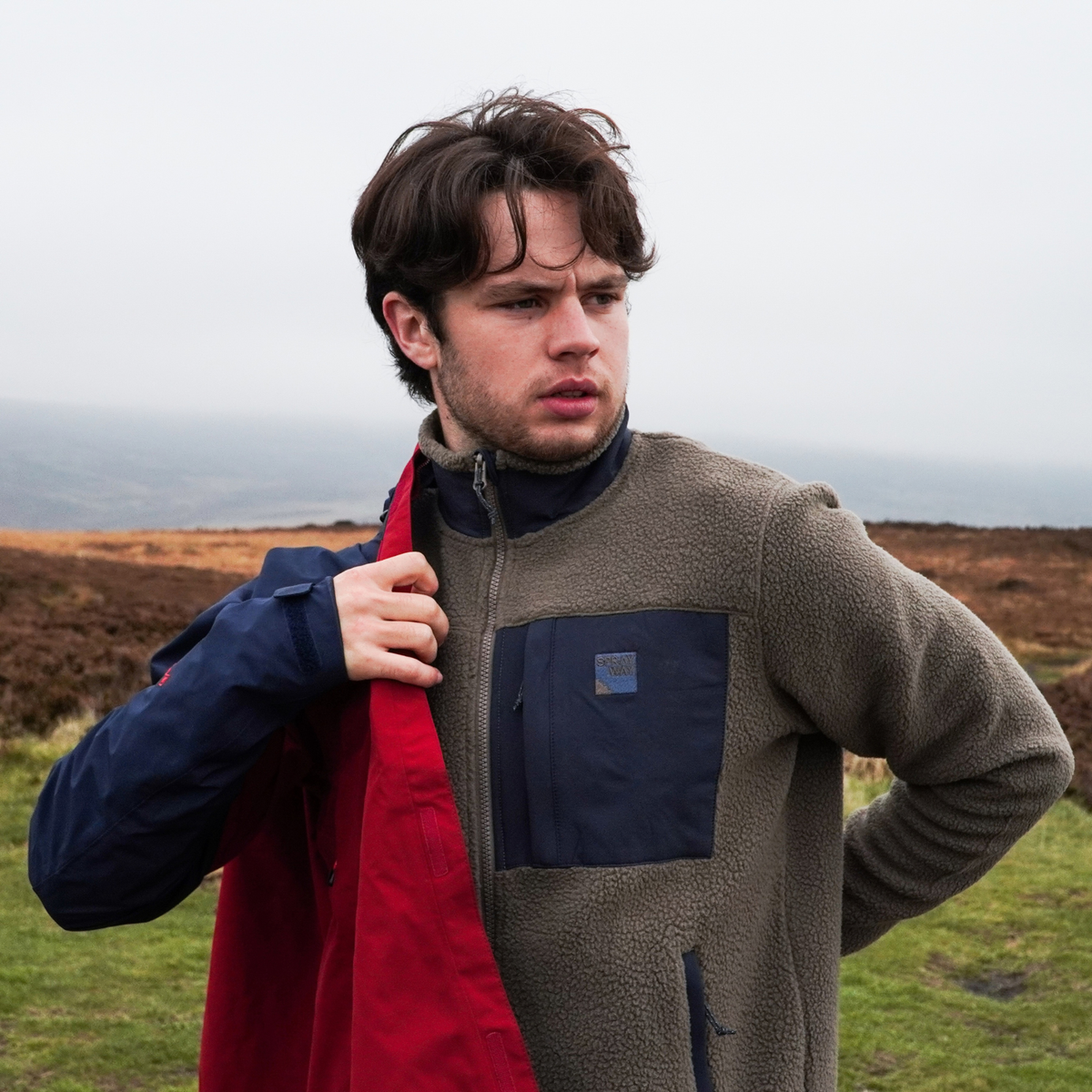A man outdoors in a grassy, hazy landscape wearing a Sprayway Men's Whitle Full Zip Fleece Jacket  SP-01737 Brownstone DkGraphite SP-007561 and putting on a red jacket.