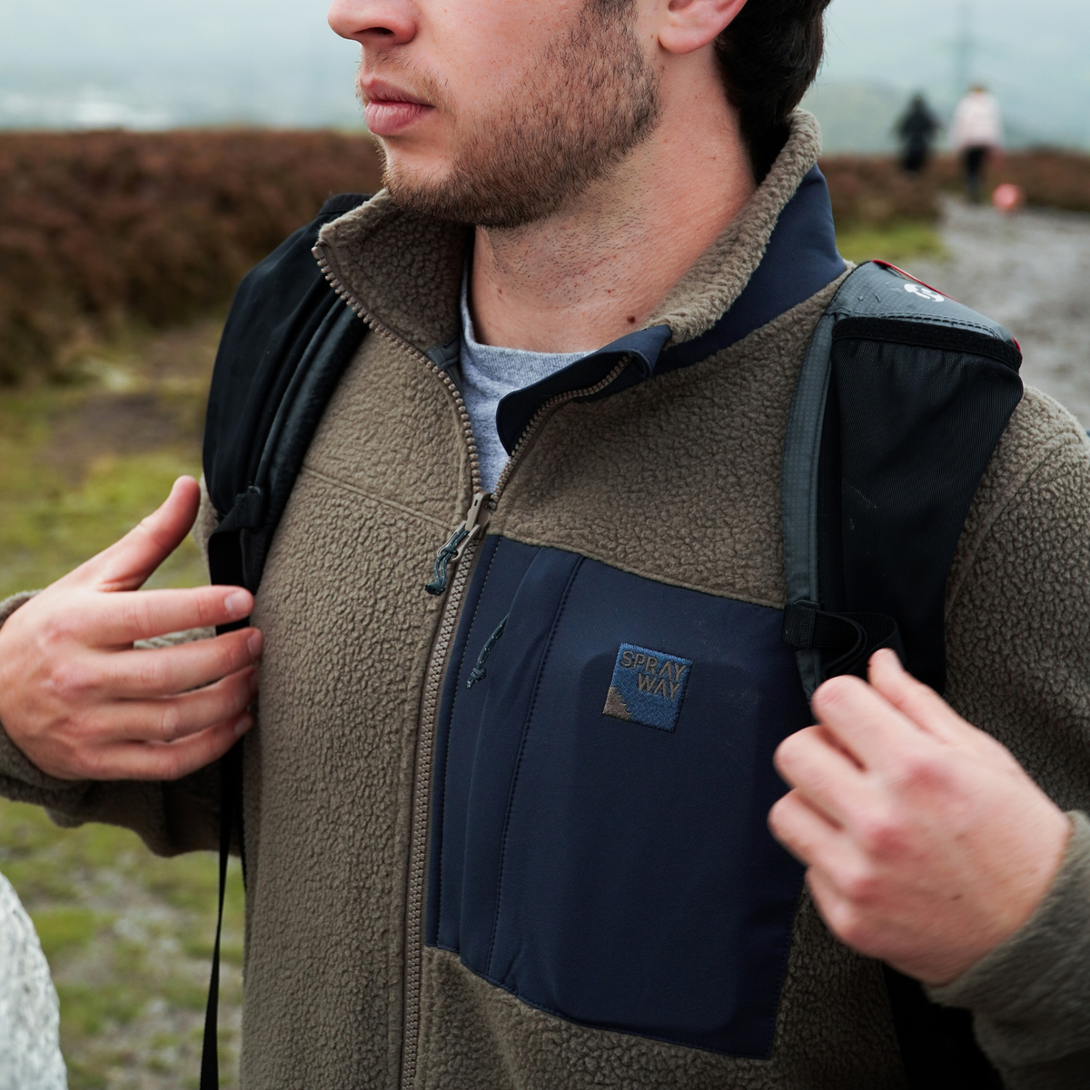 Man wearing Sprayway Men's Whitle Full Zip Fleece Jacket  SP-01737 Brownstone DkGraphite SP-007561 putting on a backpack outdoors on a mountain trail