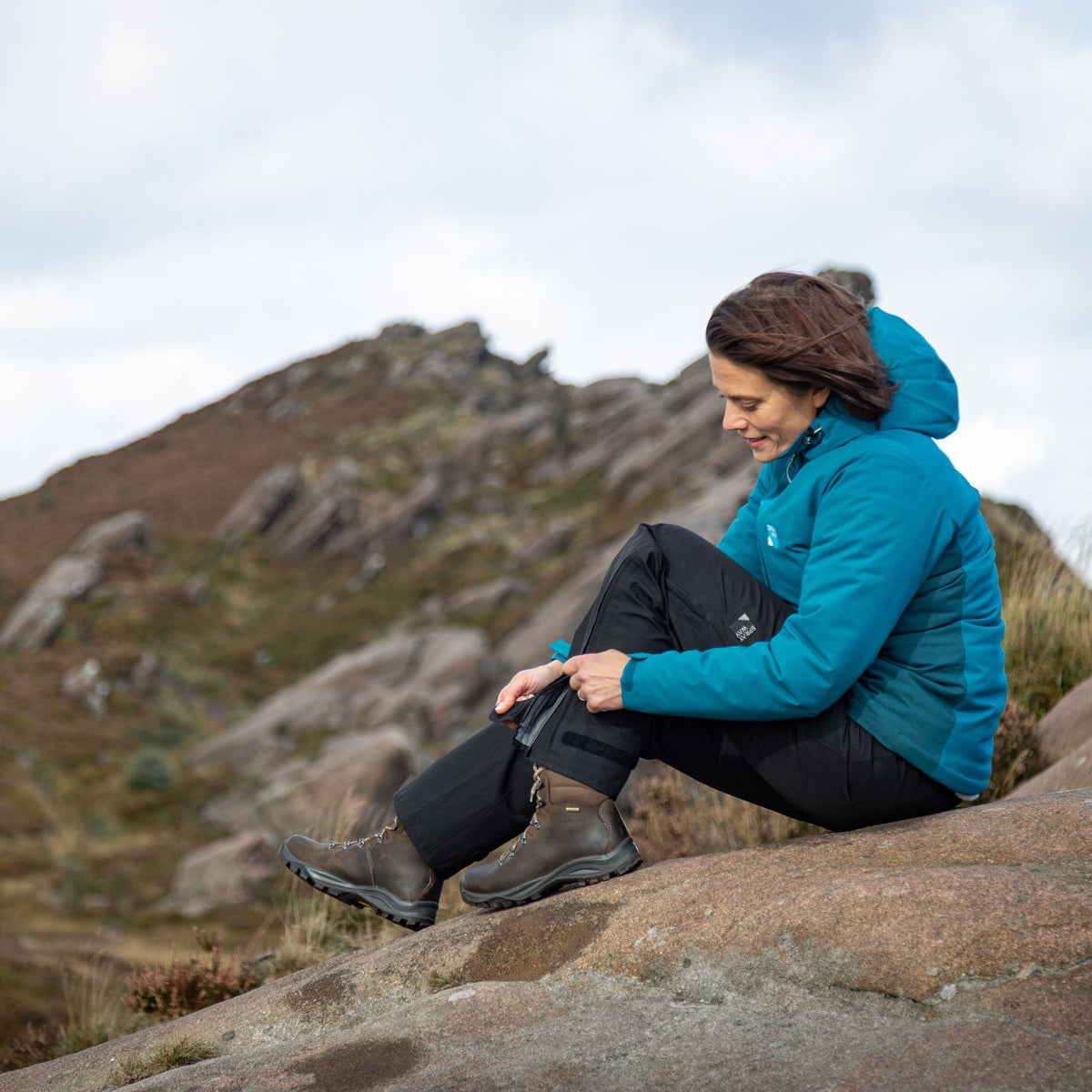 Woman wearing Sprayway Women's Mountain 3L Gore-Tex Waterproof Rain Trousers SP-01006 Black SP-008827, unzipping the leg, sitting on a boulder in the mountains.