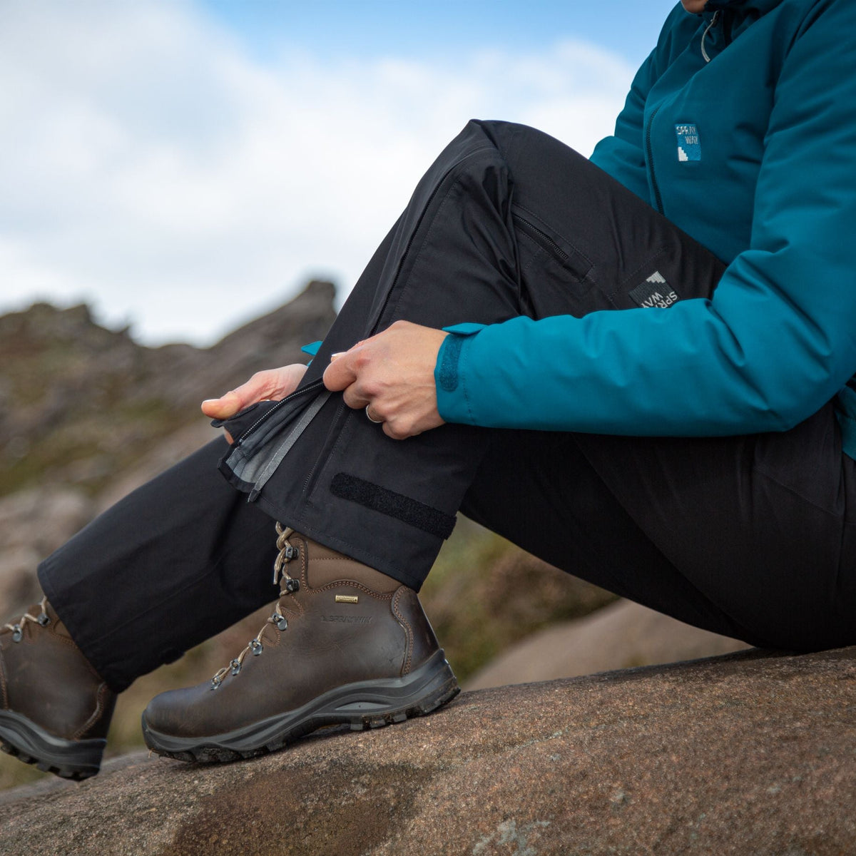 Woman wearing Sprayway Women's Mountain Gore-Tex Waterproof Rain Trousers Black SP-008827, unzipping the leg, sitting on a boulder in the mountains.