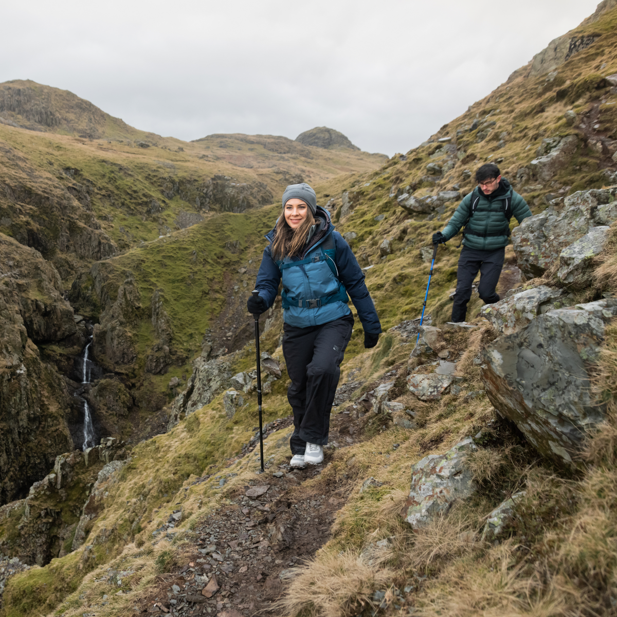 Two hikers on a rocky mountain trail with a scenic landscape. Woman using Trekmates Hiker Shock Single Pole Black TM-008705