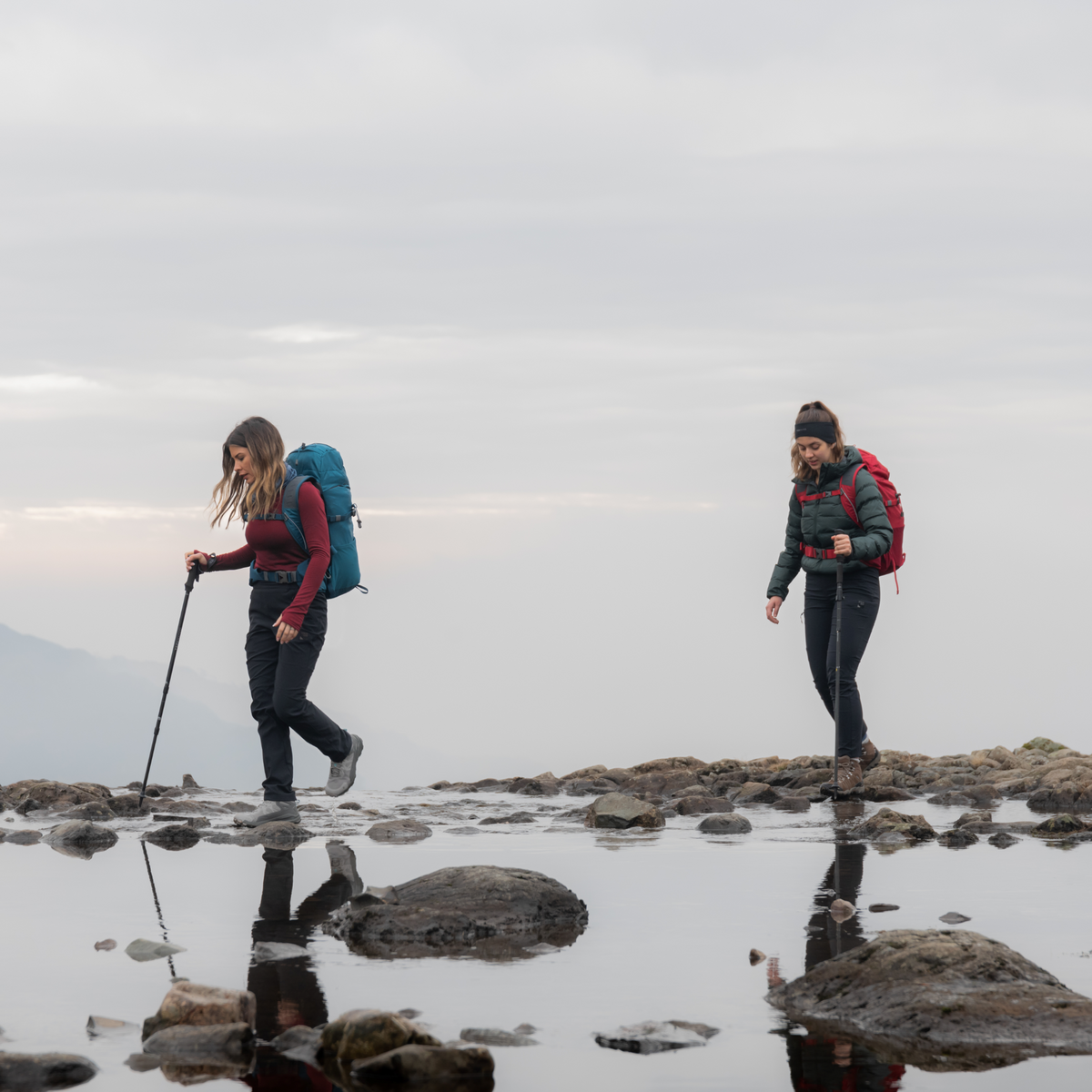 Two hikers with backpacks walking across a rocky stream with mountains in the background. One using Trekmates Hiker Shock Single Pole Black TM-008705