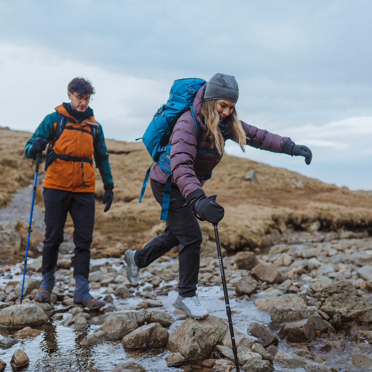 Two hikers crossing a stream in a mountainous landscape. Woman using a Trekmates Hiker Shock Single Pole Black TM-008705 to stabilize herself