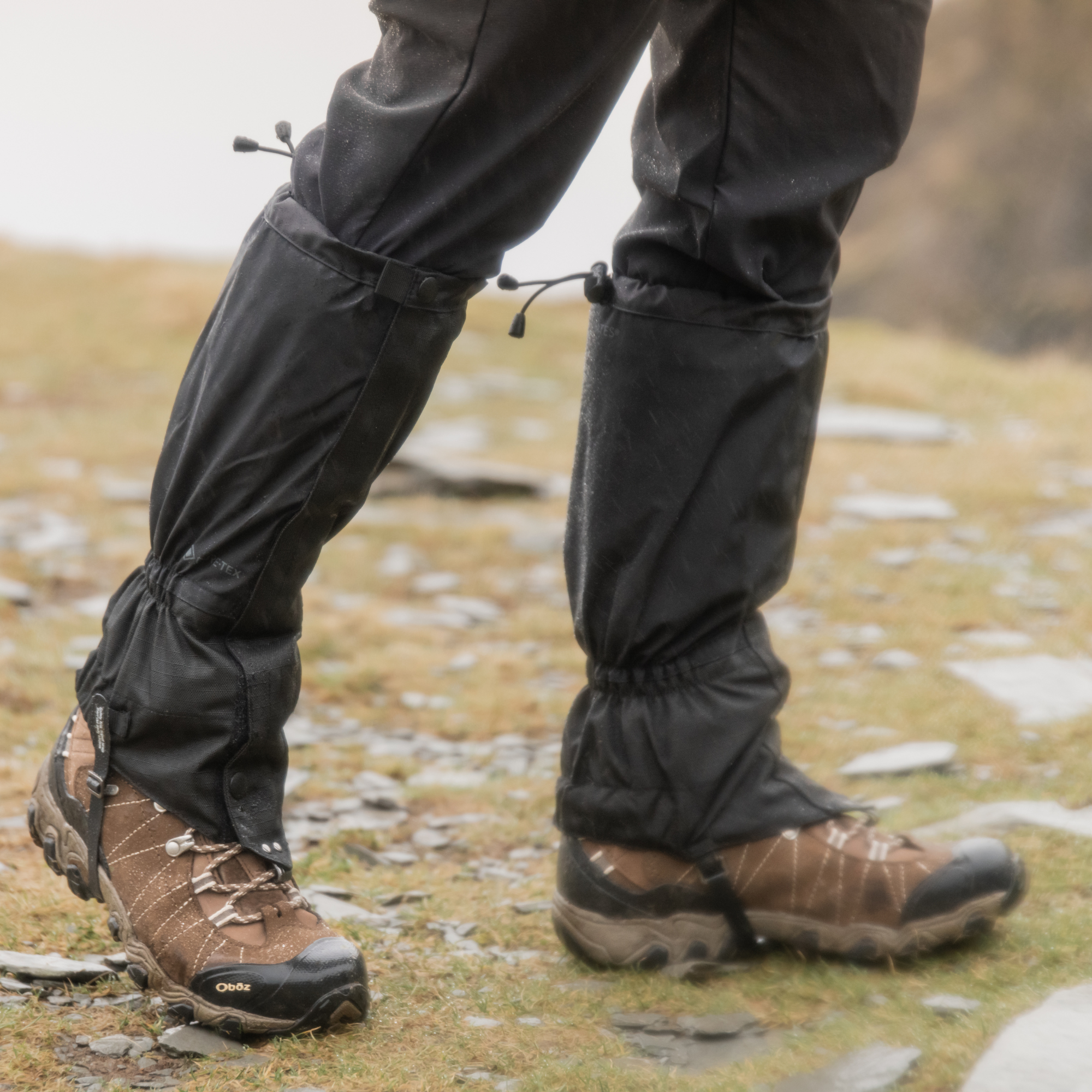 Man wearing Trekmates Unisex Cairngorm Gore-Tex Gaiters Black TM-006295 attached to black hiking trousers, worn with brown and black hiking boots on a rocky, grassy outdoor trail.