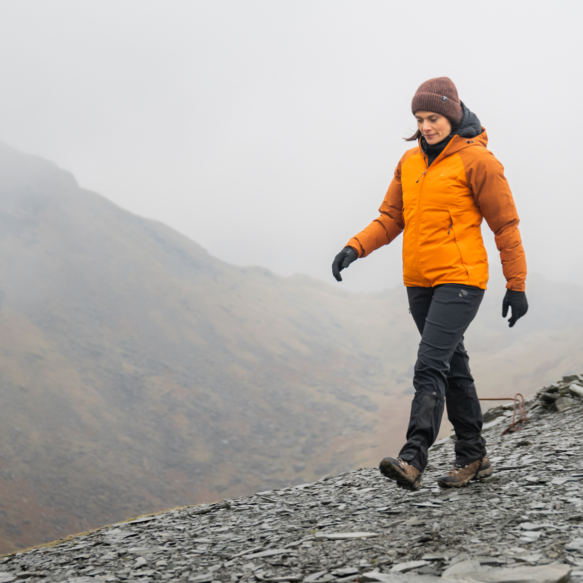 A woman hiking on a rocky mountain path wearing an orange hooded jacket, dark grey trousers with Trekmates Unisex Cairngorm Gore-Tex Gaiters Black TM-006295 attached, black gloves, and a brown beanie, with a misty mountain landscape in the background.