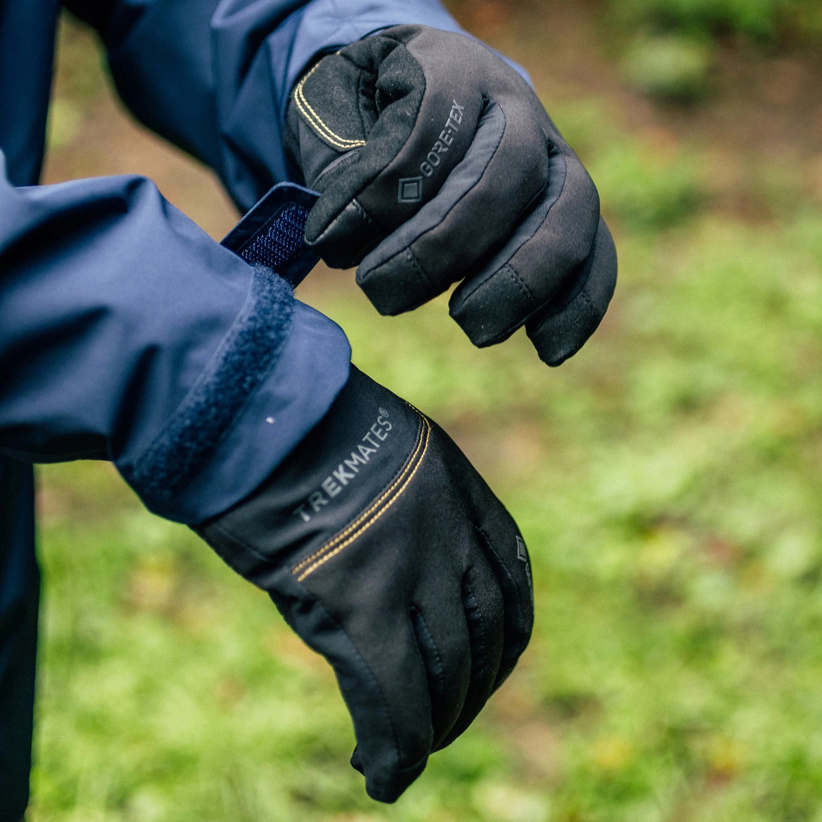 A close-up of a person wearing Trekmates Unisex Friktion Gore-Tex Waterproof Gloves Black TM-008647 with yellow stitching, shown outdoors against a blurred green background.