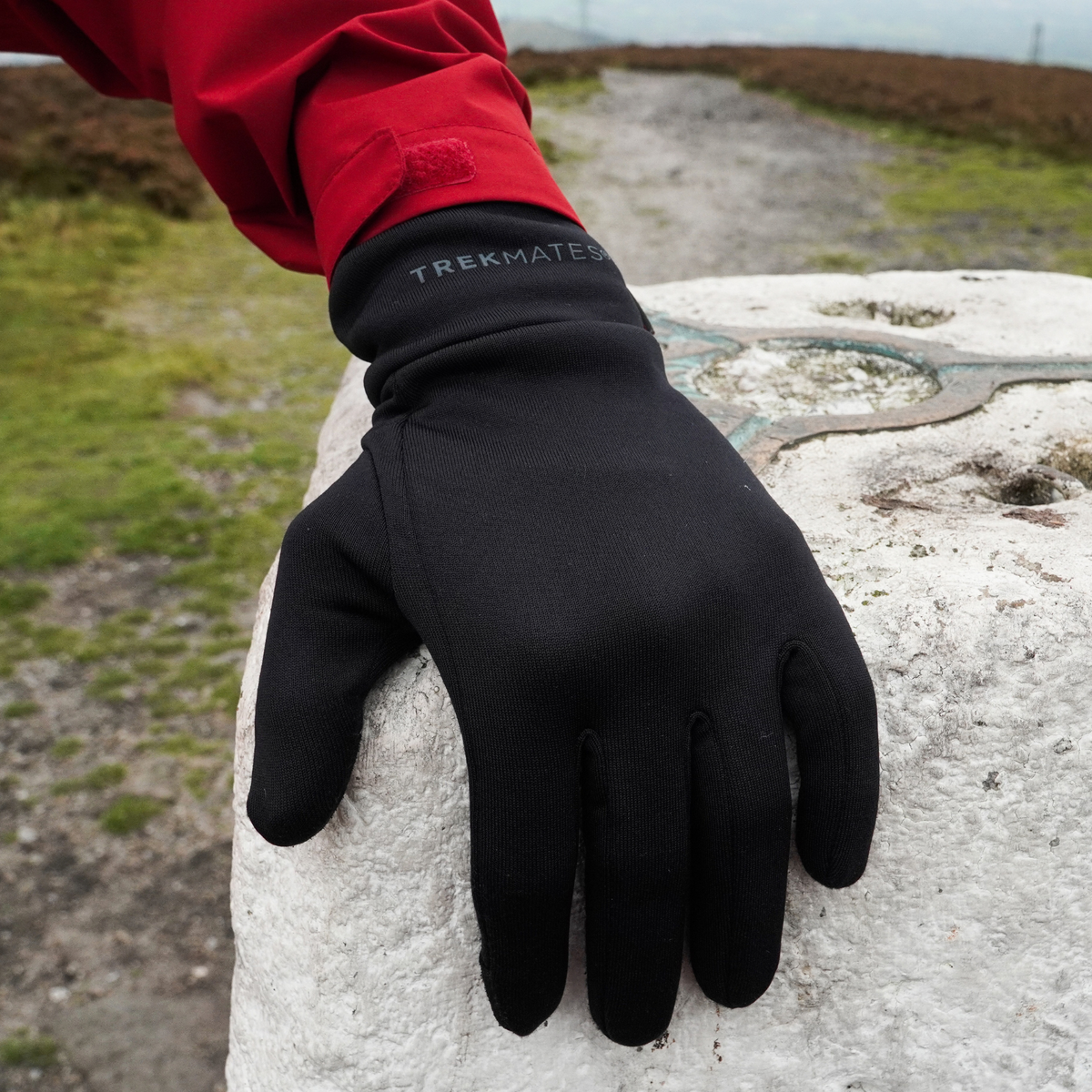 A man wearing Trekmates Unisex Silk Touch Liner Gloves Black GLV-TH-U10547 with his hand placed on a summit marker on a trail