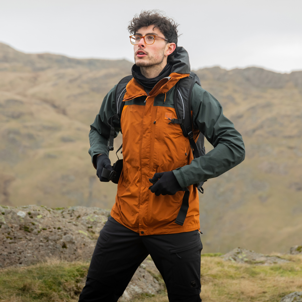 Man wearing a waterproof jacket and Trekmates Unisex Silk Touch Liner Gloves Black GLV-TH-U10547 clipping a backpack strap with a mountain background.