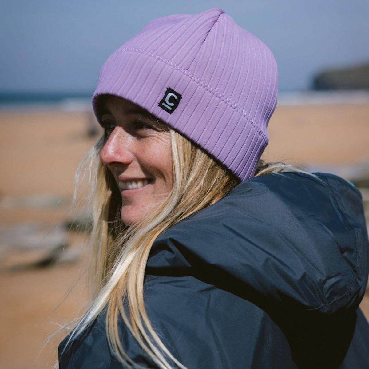 Woman wearing a Unisex Storm Chaser Thermal Neoprene Swim Watersports Beanie Lilac C-HOBE and a hardshell changing robe on a beach
