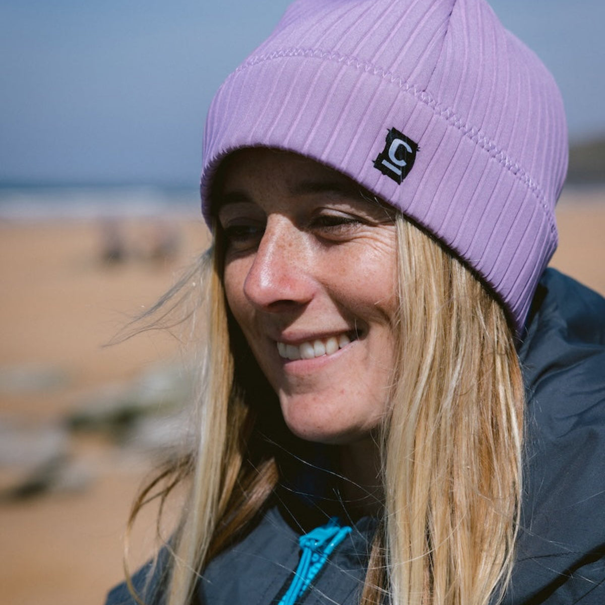 Woman wearing a Unisex Storm Chaser Thermal Neoprene Swim Watersports Beanie Lilac C-HOBE on a beach with a hardshell changing robe