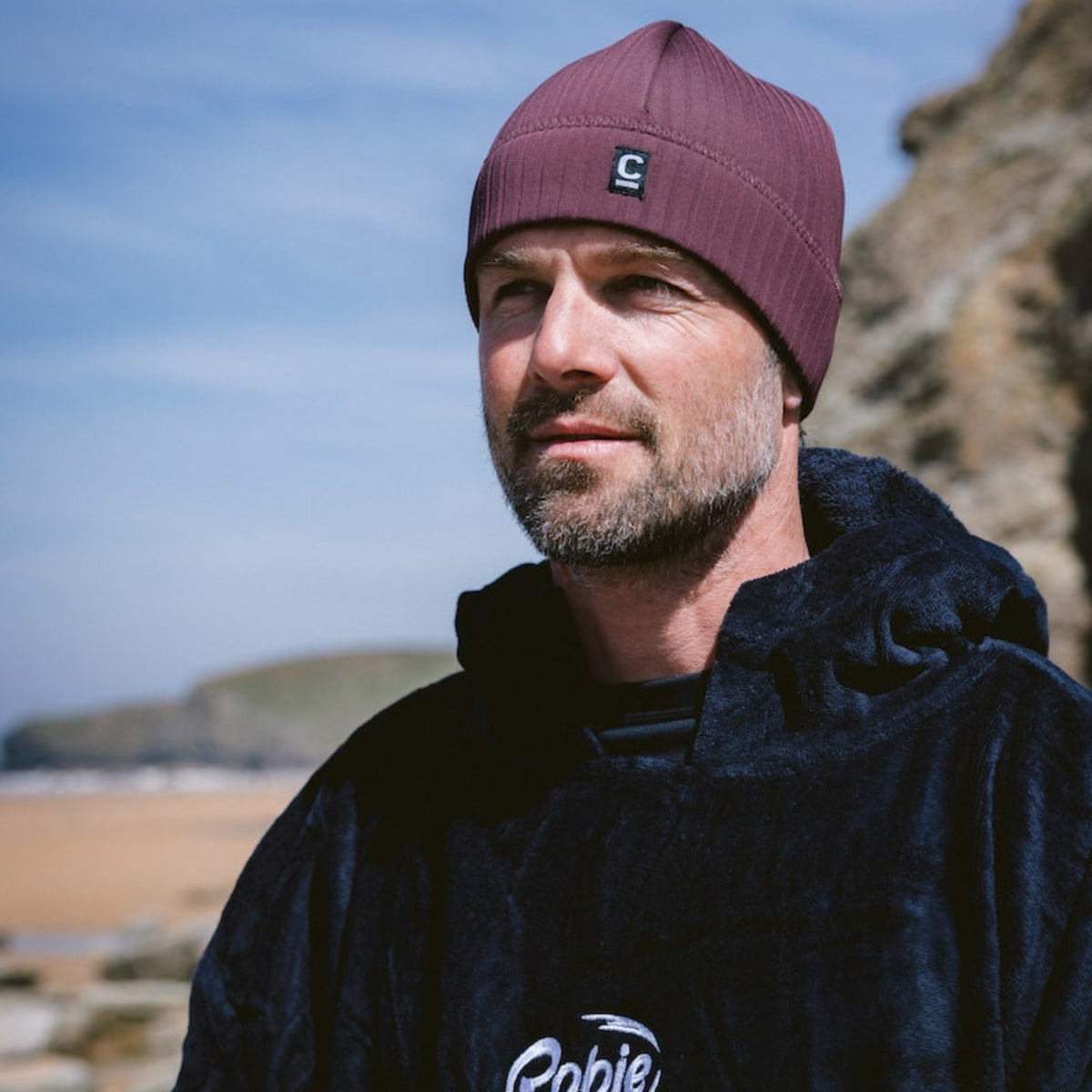 Man wearing a C-Skins Unisex Storm Chaser Thermal Neoprene Swim Watersports Beanie Rust C-HOBE and black Robie changing robe, standing outdoors against a blue sky.