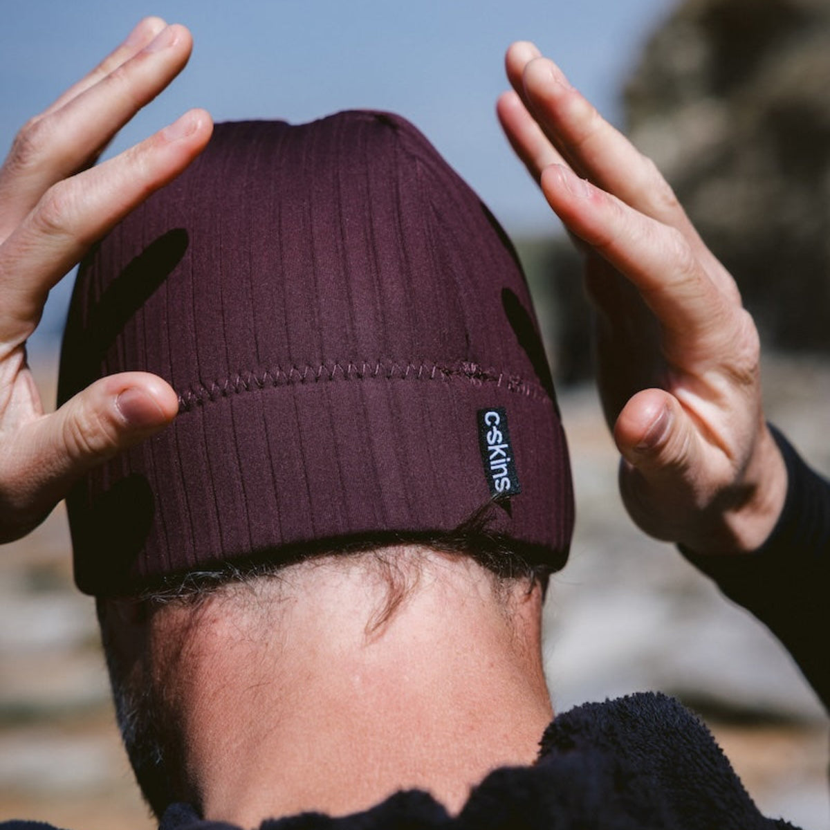 Man wearing a C-Skins Unisex Storm Chaser Thermal Neoprene Swim Watersports Beanie Rust C-HOBE and black Robie changing robe, standing outdoors against a blue sky.
