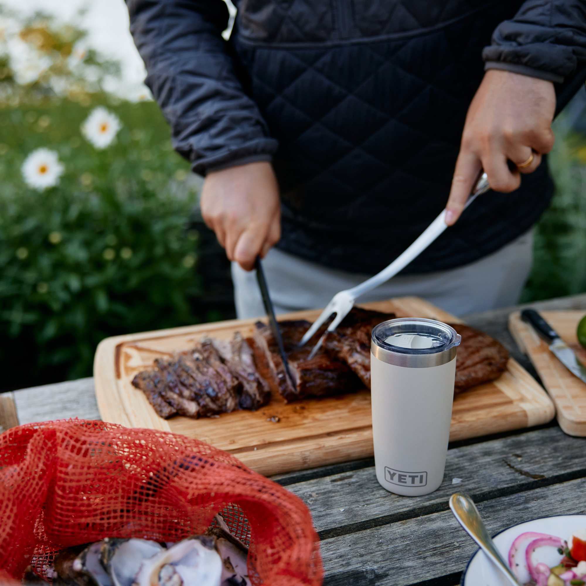 A person outdoors at a wooden table carving a steak on a cutting board, with a Yeti Rambler 10oz Tumbler Cape Taupe 70000003165 Yeti-RamblerTumbler-10oz on the table and a red mesh bag in the foreground.