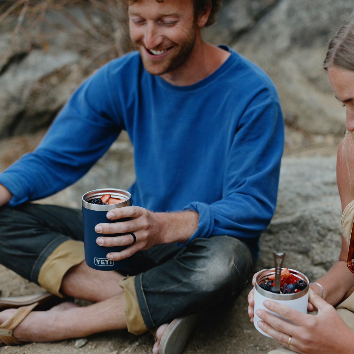 A man in a blue long-sleeve shirt sits outdoors on rocks, smiling while holding a navy blue Yeti Rambler insulated food jar. A woman is partially visible on the right holding a white Yeti food jar with berries.