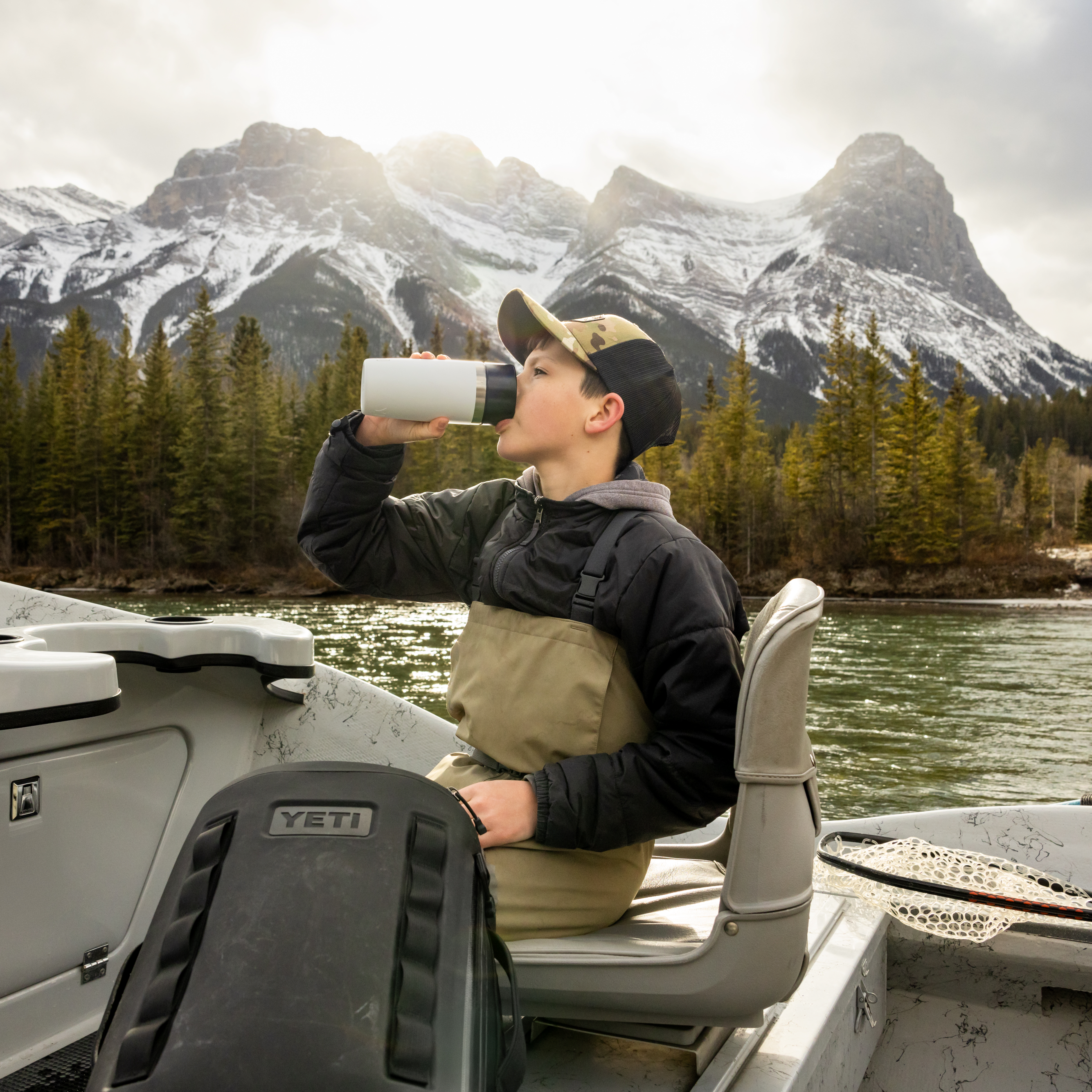 Young boy drinking out of a white Yeti travel bottle with commuter lid on a fishing boat wearing fishing waders