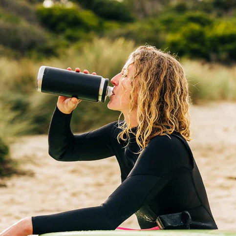Woman drinking out of a Yeti Rambler 26oz Chug Cap Bottle Yeti-Chug-26oz in a wetsuit on the beach