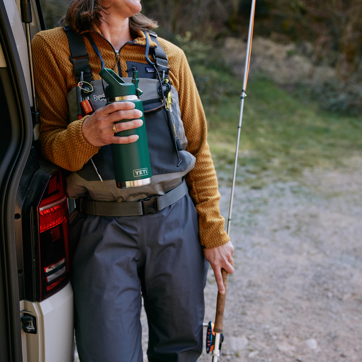 A person outdoors by the open rear door of a vehicle, wearing a brown knit long-sleeve top, grey waders, and a dark grey chest rig. They are holding a Yeti Rambler 26oz Insulated Bottle Straw Cap Black Forest Green 70000004236 Yeti-RamblerStraw-26oz and holding a fishing rod.