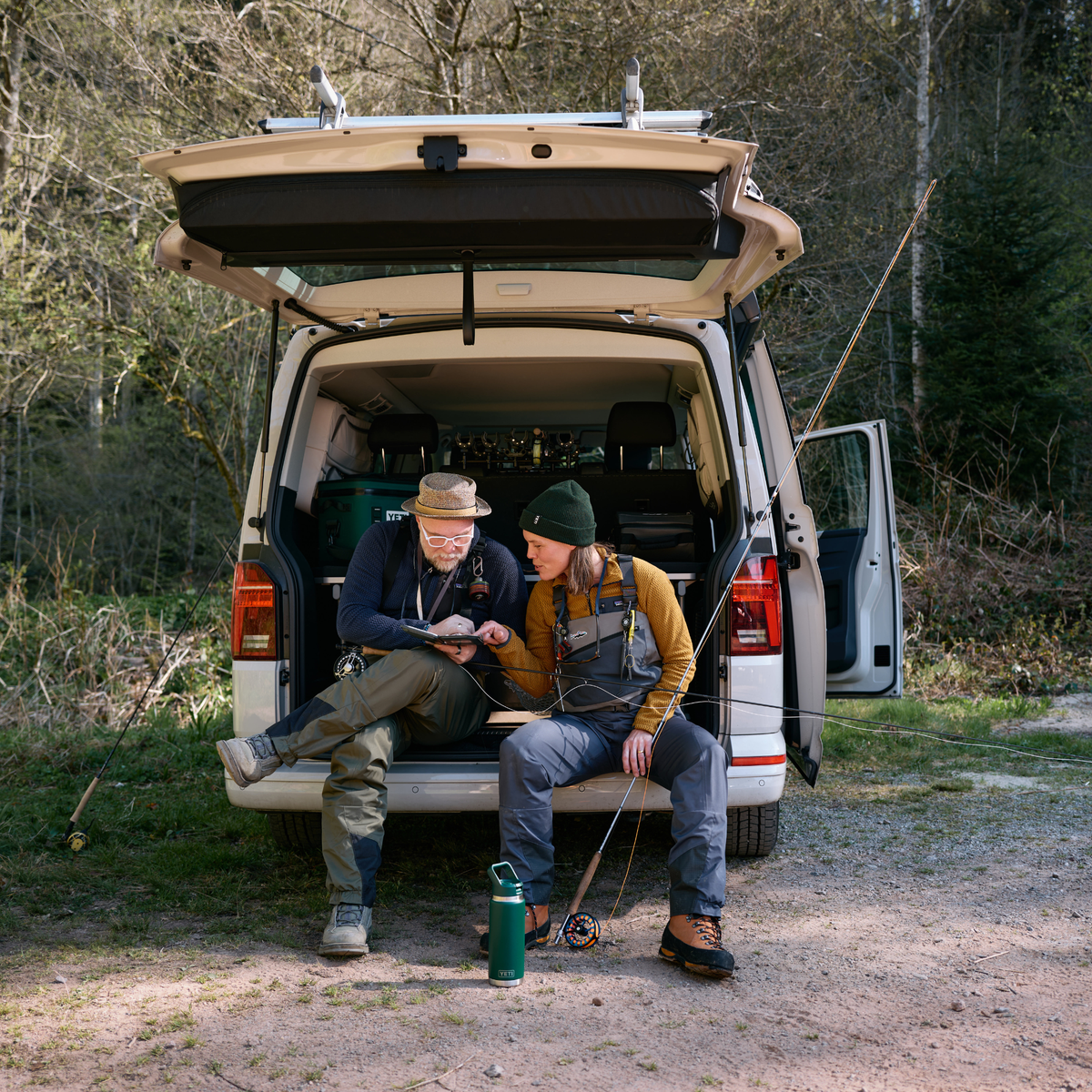 Two people sit on the open boot of a beige van in a wooded outdoor setting, fishing with rods and line. The van is parked on a dirt path with trees and grass in the background. A Yeti Rambler 26oz Insulated Bottle Straw Cap Black Forest Green 70000004236 Yeti-RamblerStraw-26oz sits on the ground in front of them.
