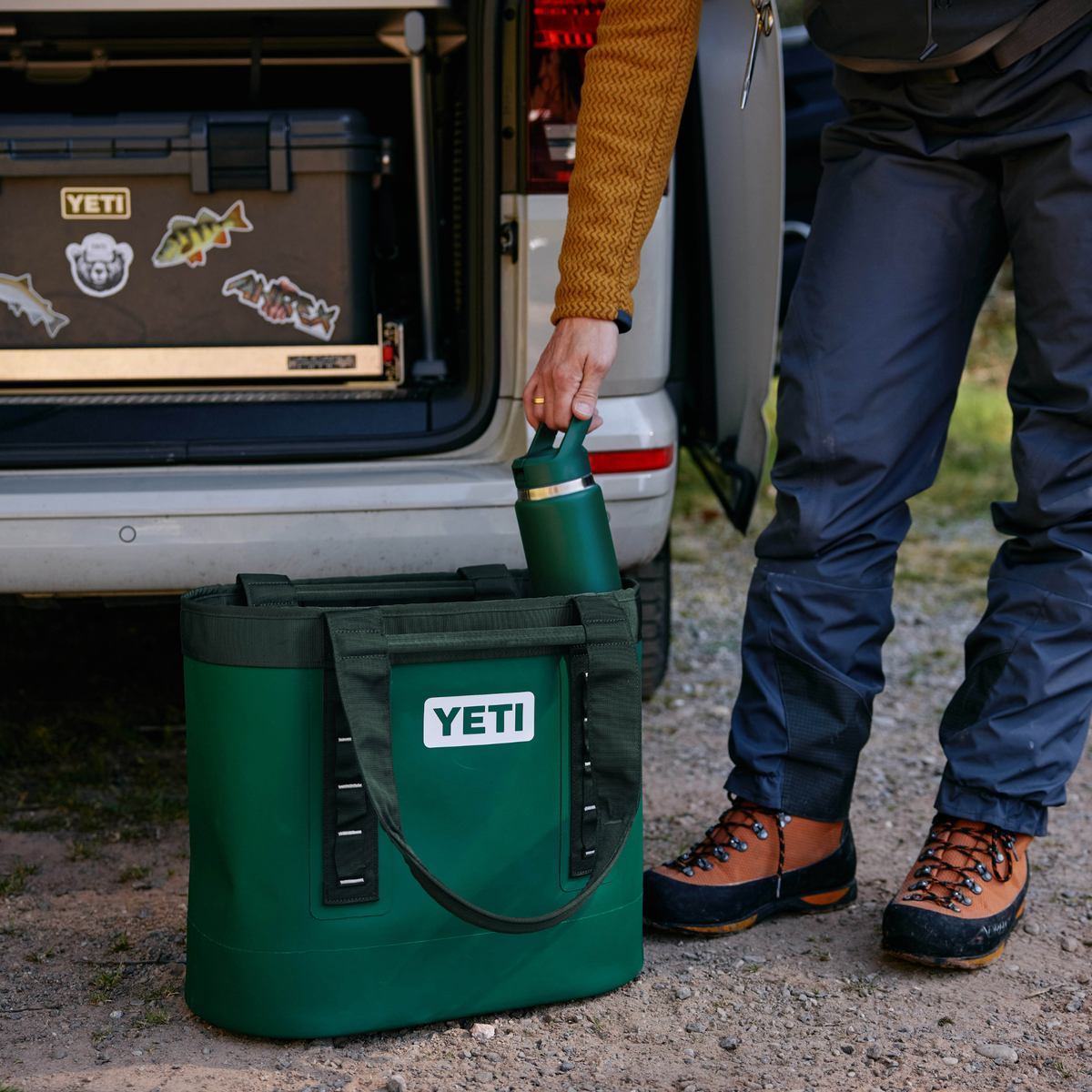 A green YETI cooler bag sits on gravel next to an open vehicle boot, with a person reaching in to place a matching Yeti Rambler 26oz Insulated Bottle Straw Cap Black Forest Green 70000004236 Yeti-RamblerStraw-26oz into the bag.