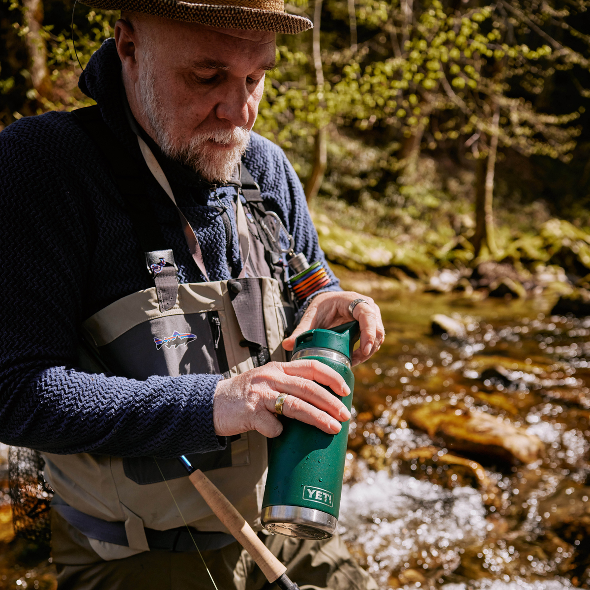 A man in outdoor fishing attire stands by a flowing stream, holding a Yeti Rambler 26oz Insulated Bottle Straw Cap Black Forest Green 70000004236 Yeti-RamblerStraw-26oz. He wears a brown hat, a dark blue knit sweater, and a grey fishing vest, with a blurred forest and stream in the background.