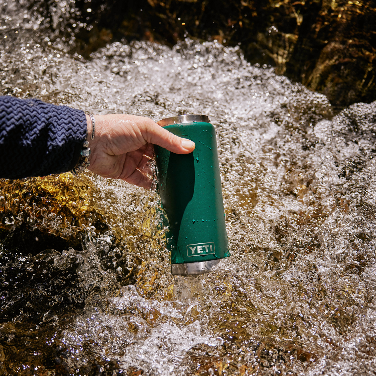 A hand wearing a navy blue knit sleeve holds a Yeti Rambler 26oz Insulated Bottle Straw Cap Black Forest Green 70000004236 Yeti-RamblerStraw-26oz above rushing water outdoors.