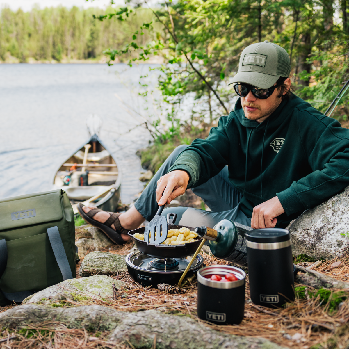 A man wearing a green hoodie, green cap, and sunglasses sits by a lake outdoors, cooking food in a small pan on a portable burner. A green bag and 2 black Yeti Rambler insulated food jars are placed nearby on rocks with pine needles and trees in the background.