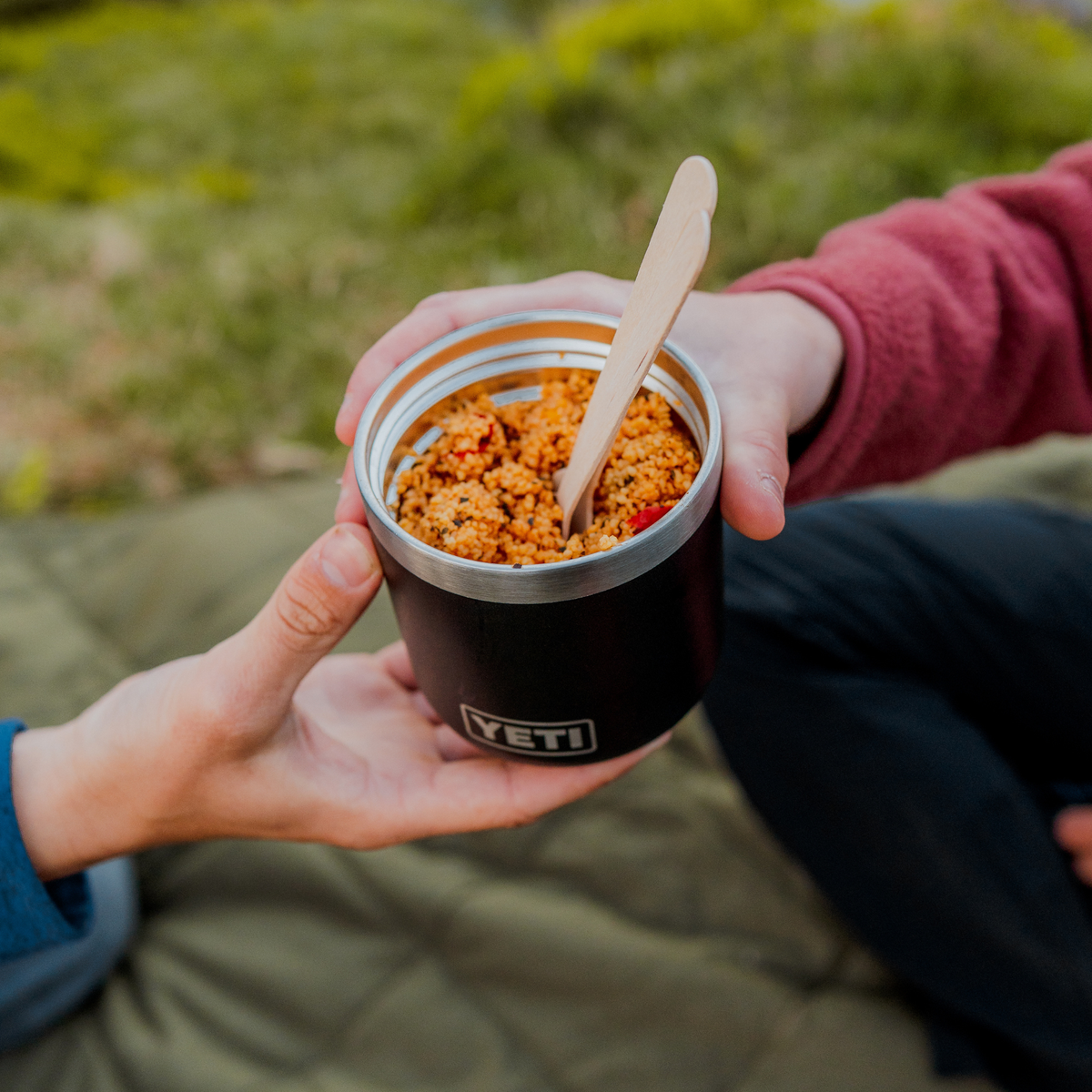 A close-up lifestyle shot of a person holding a Yeti Rambler insulated food jar filled with a orange-brown food, with a wooden spoon inside.